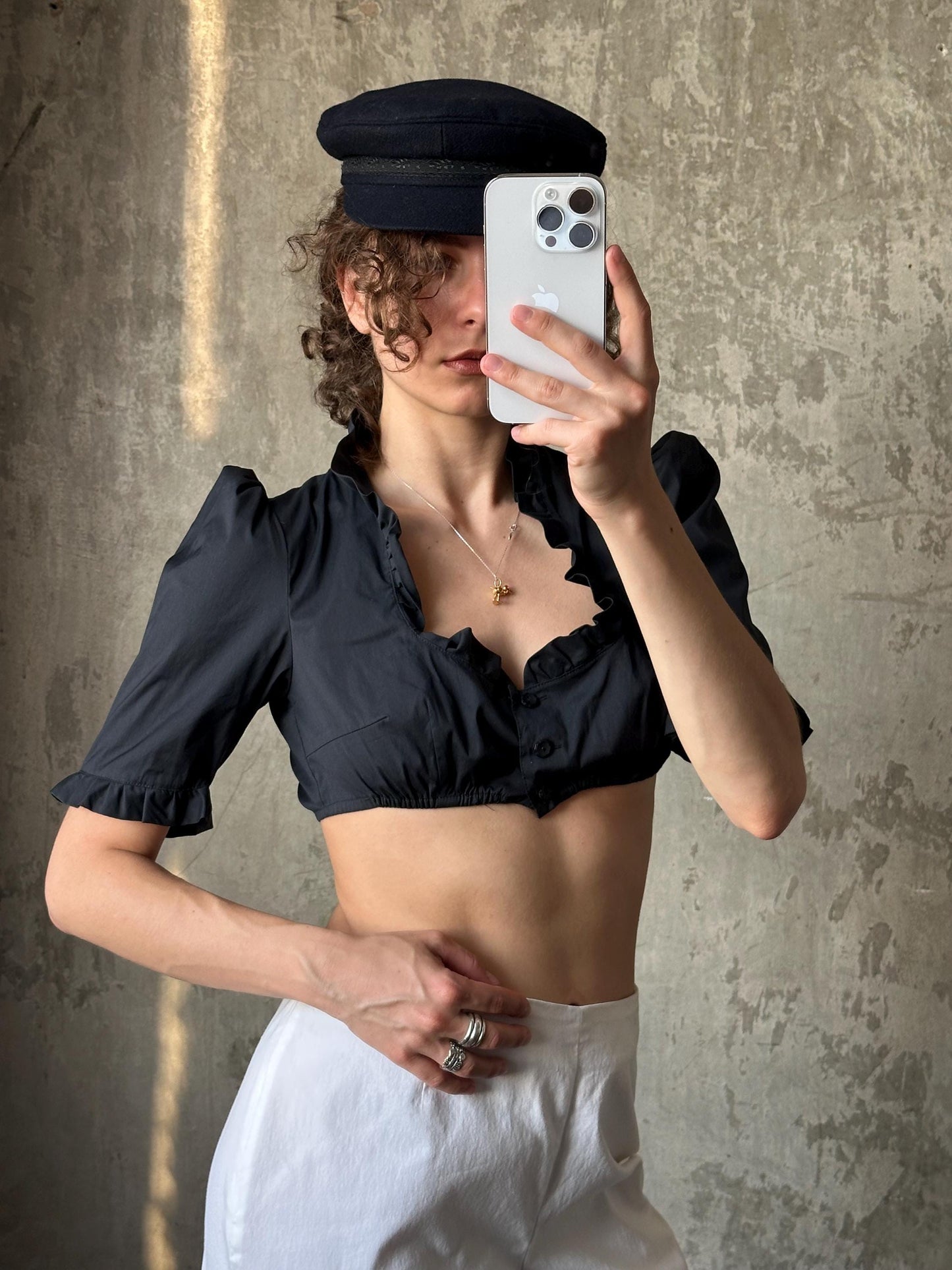 A woman is taking a selfie with her iPhone, wearing a black crop top and a black hat.