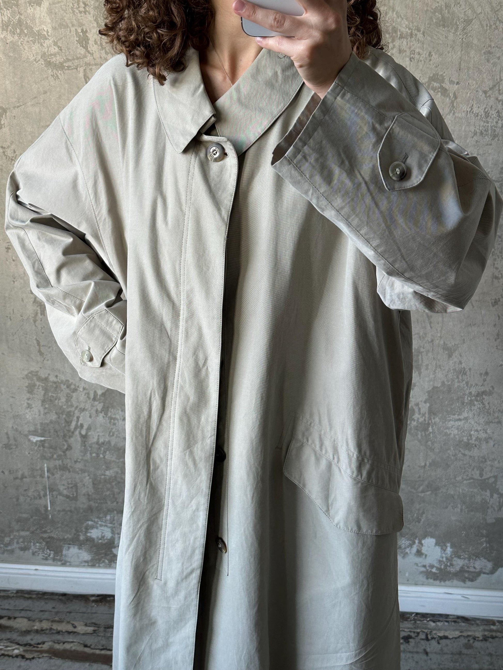A person is taking a selfie with a camera, wearing a light-colored coat.