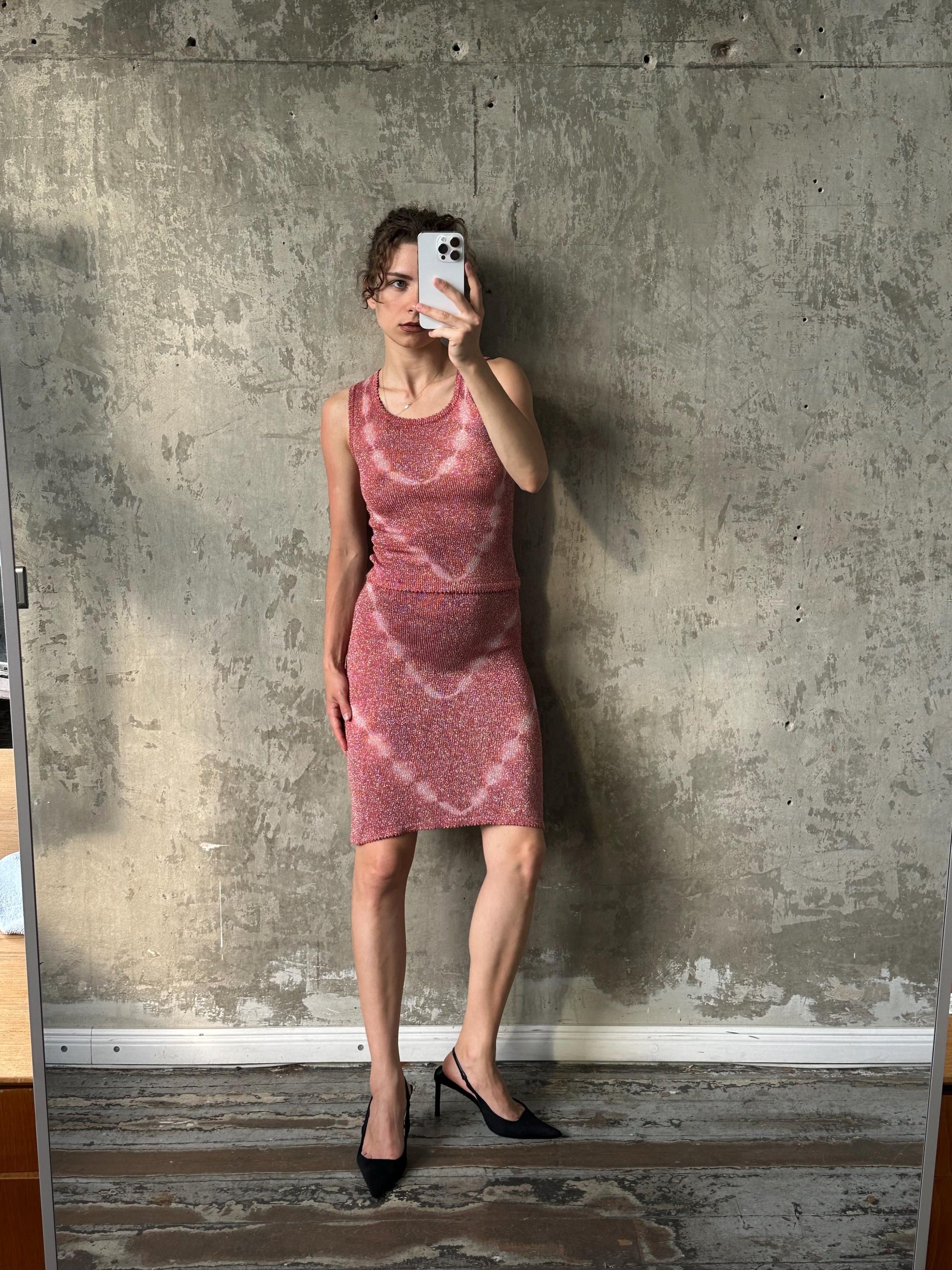 A woman in a pink dress is taking a selfie in front of a concrete wall.