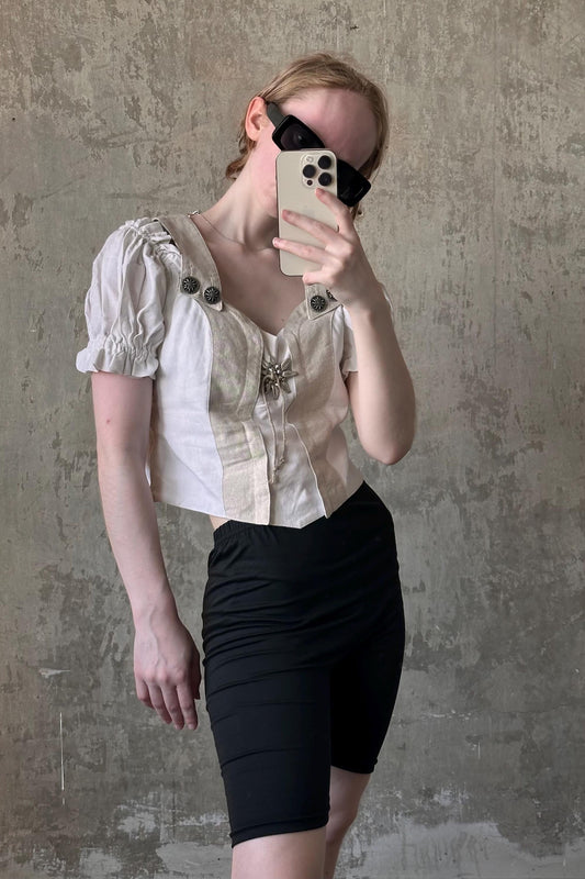 A person is taking a selfie with a smartphone, wearing a white blouse with puffed sleeves and black shorts.