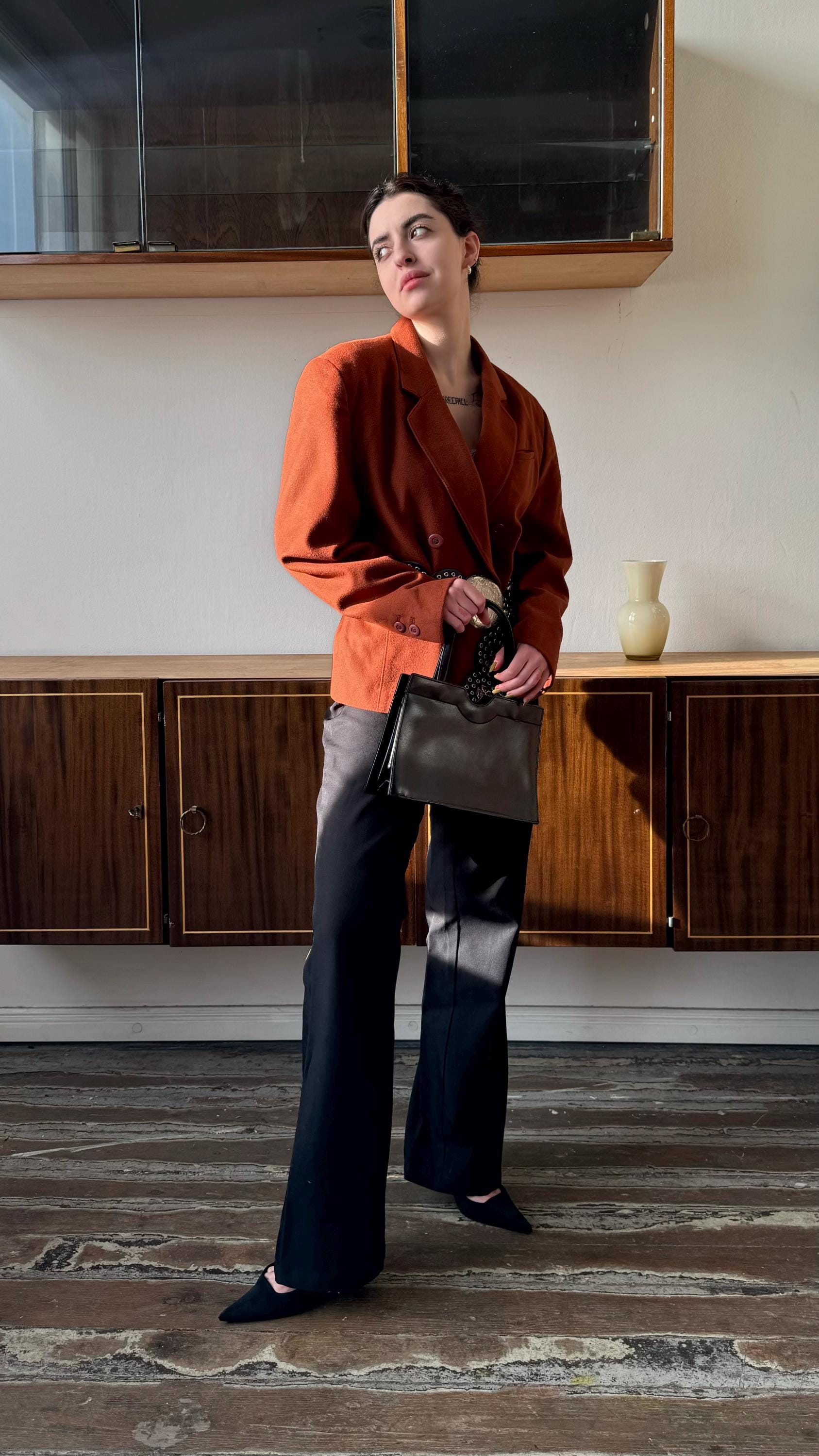 A woman in a red blazer and black pants stands in a room with wooden cabinets, holding a small bag.