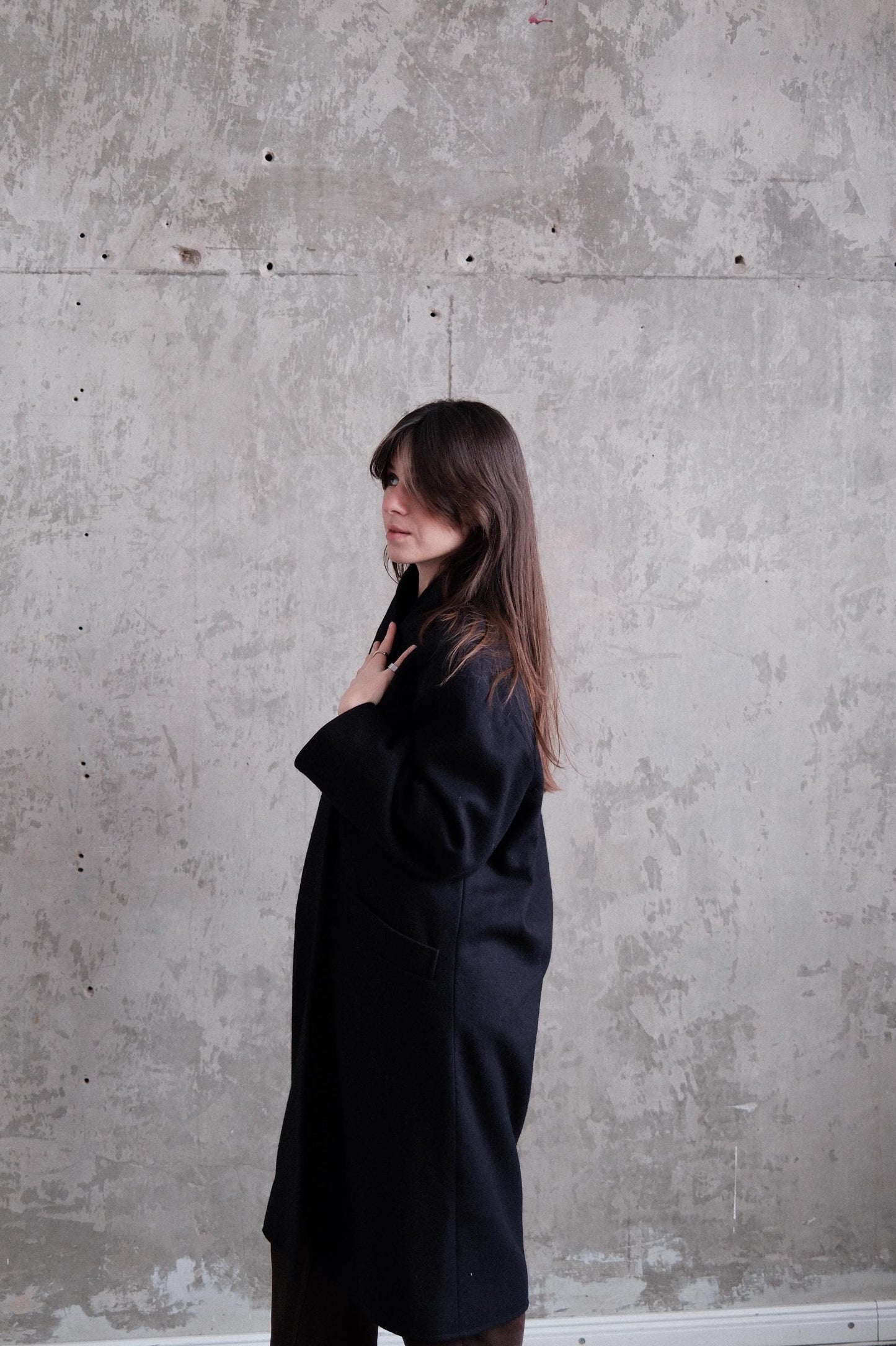 A woman in a black coat stands against a textured concrete wall, her expression conveying a sense of contemplation or introspection.