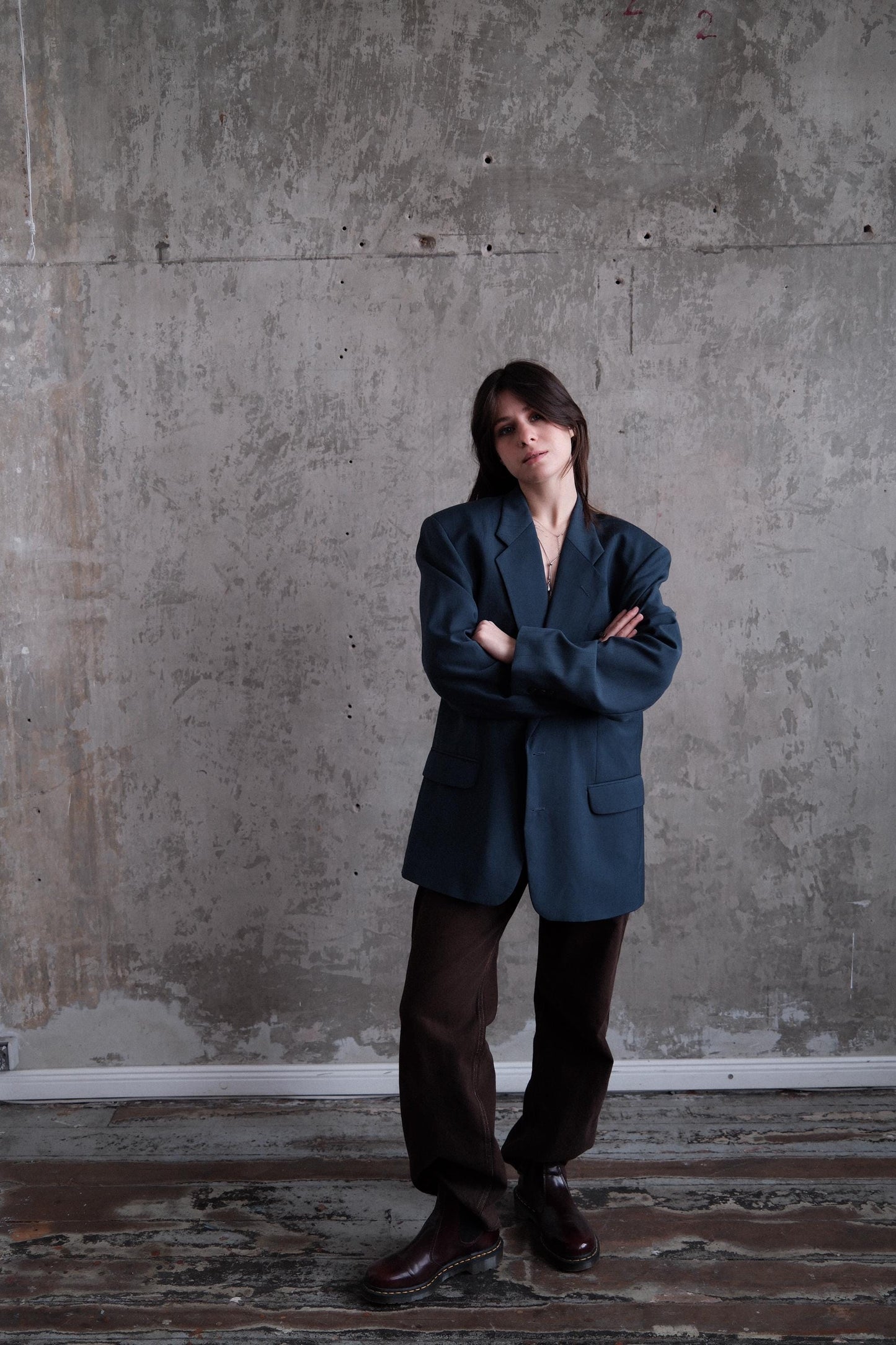 A person wearing a dark blue blazer and brown pants stands with their arms crossed against a concrete wall.