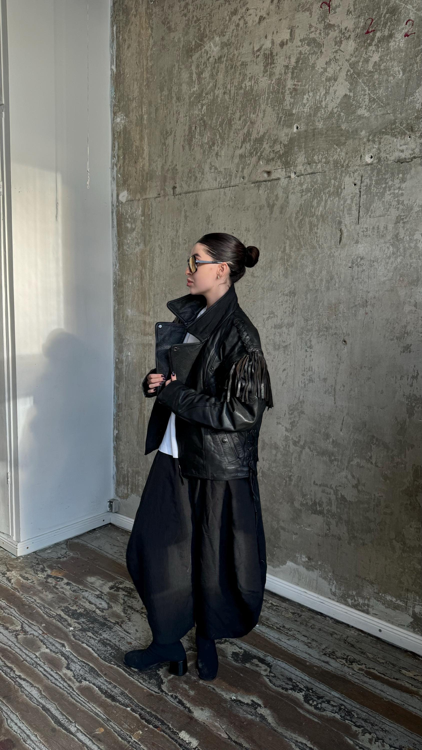 A woman wearing a black leather jacket and sunglasses stands in a room with a concrete wall.