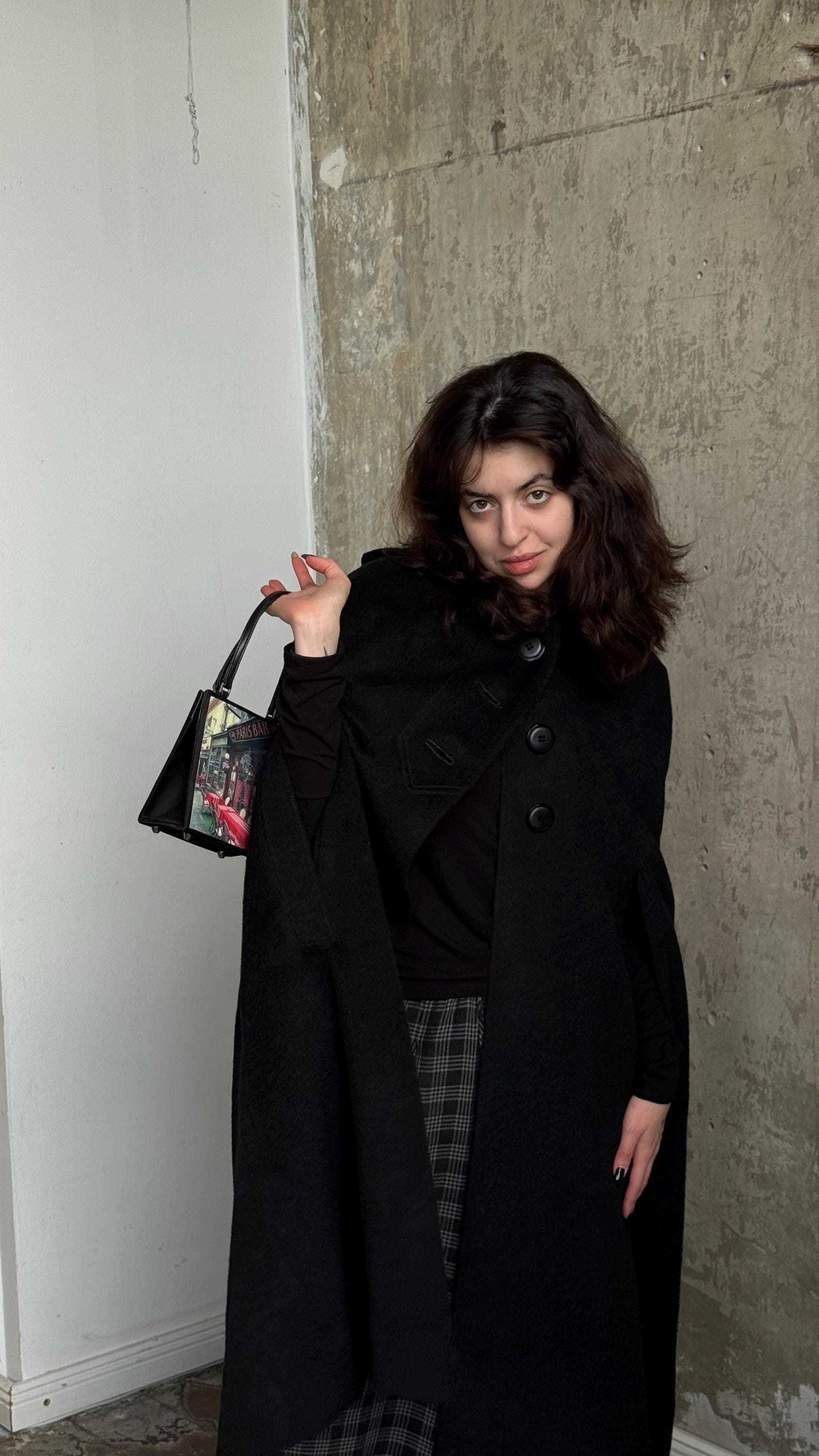 A woman in a black coat and plaid skirt stands in front of a concrete wall, holding a purse and looking directly at the camera.