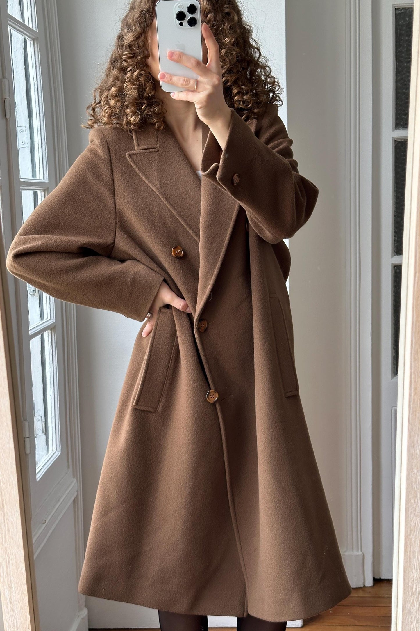 A woman with curly hair is taking a selfie in front of a mirror, wearing a brown coat.