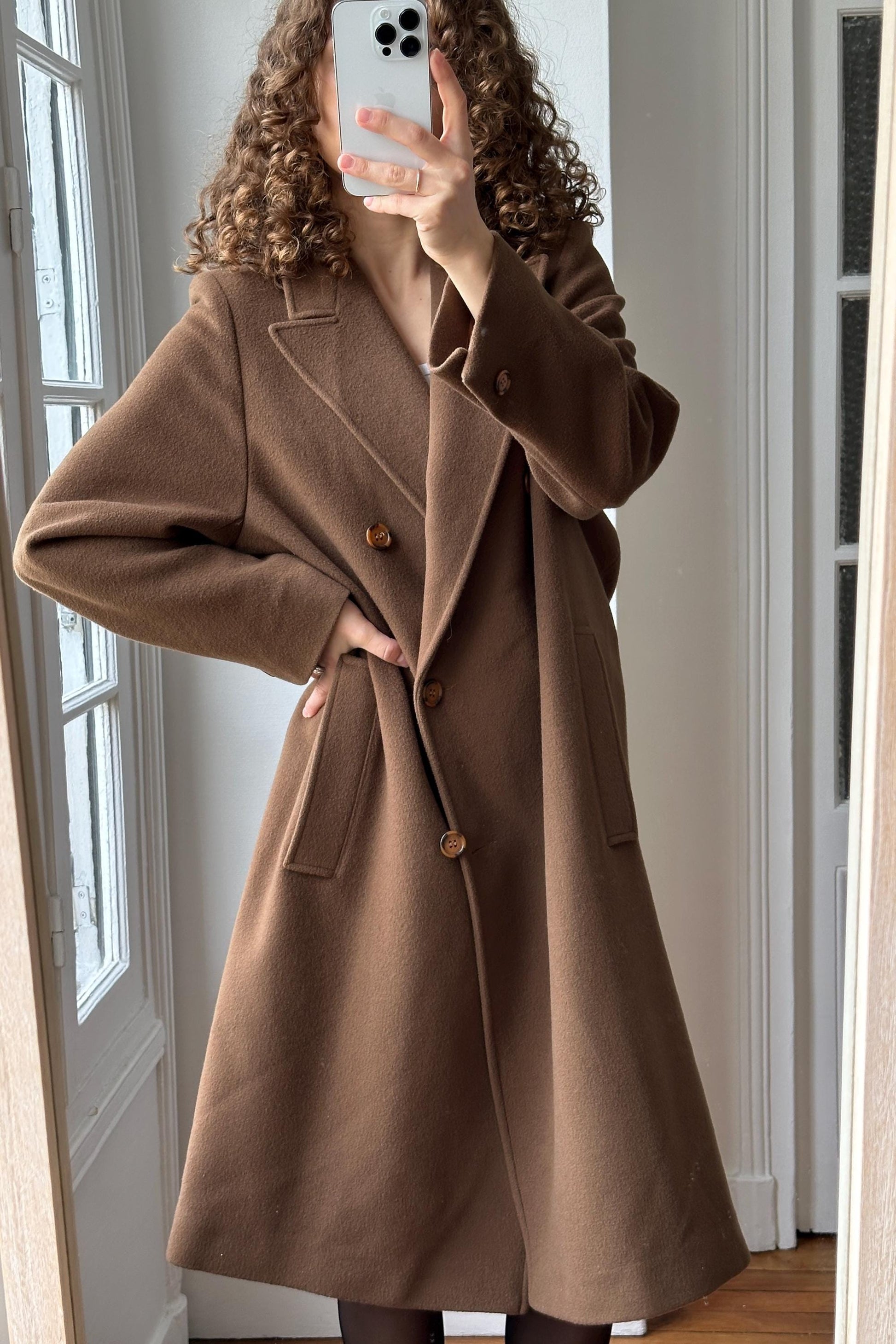 A woman with curly hair is taking a selfie in front of a mirror, wearing a brown coat.