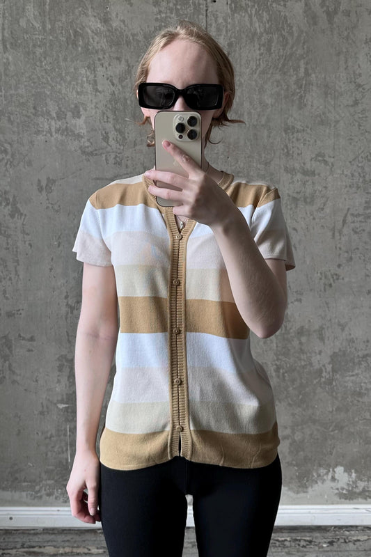 A woman wearing a striped sweater and sunglasses is taking a selfie with her iPhone against a concrete wall.