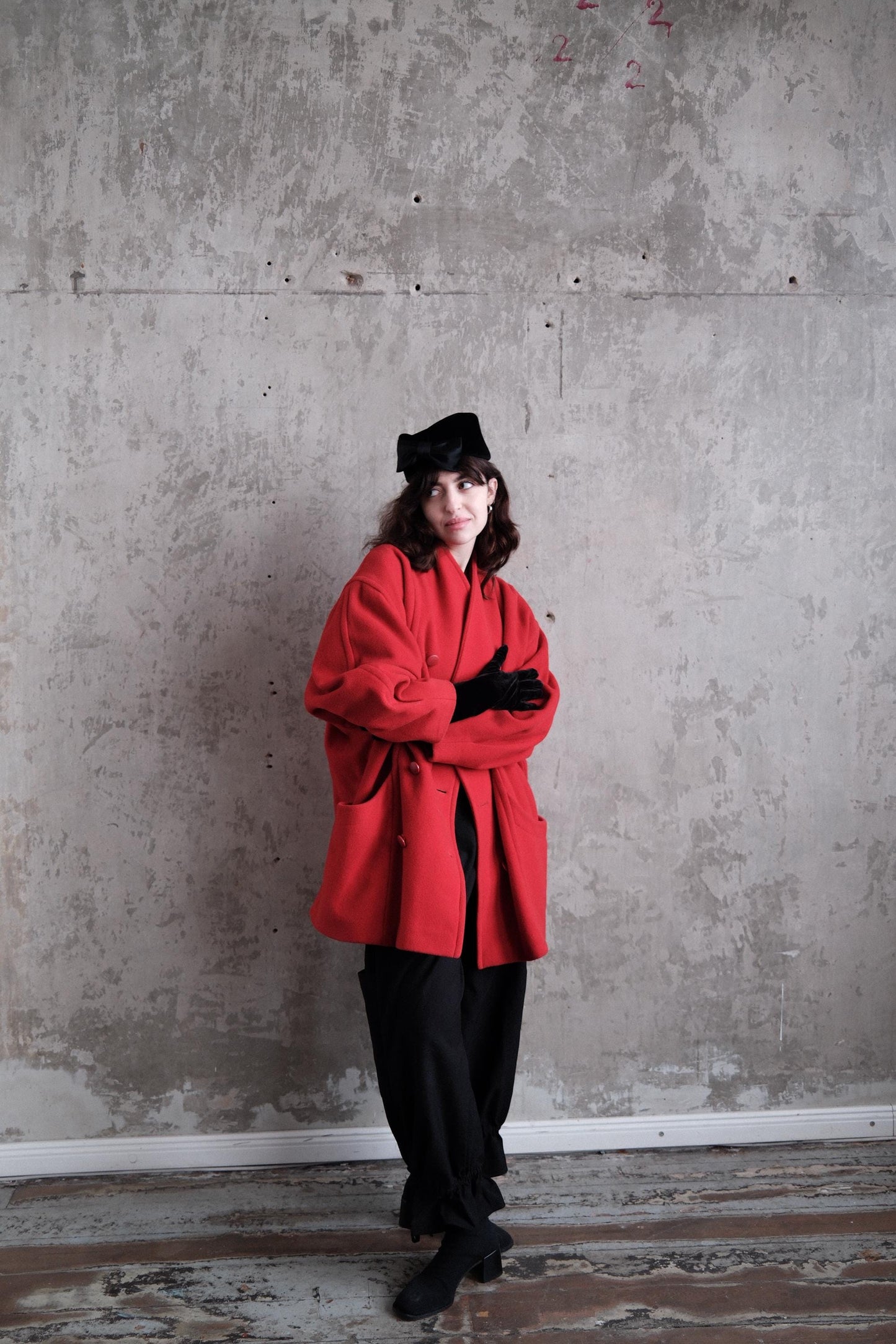 A person wearing a red coat and black pants stands in a room with a concrete wall.