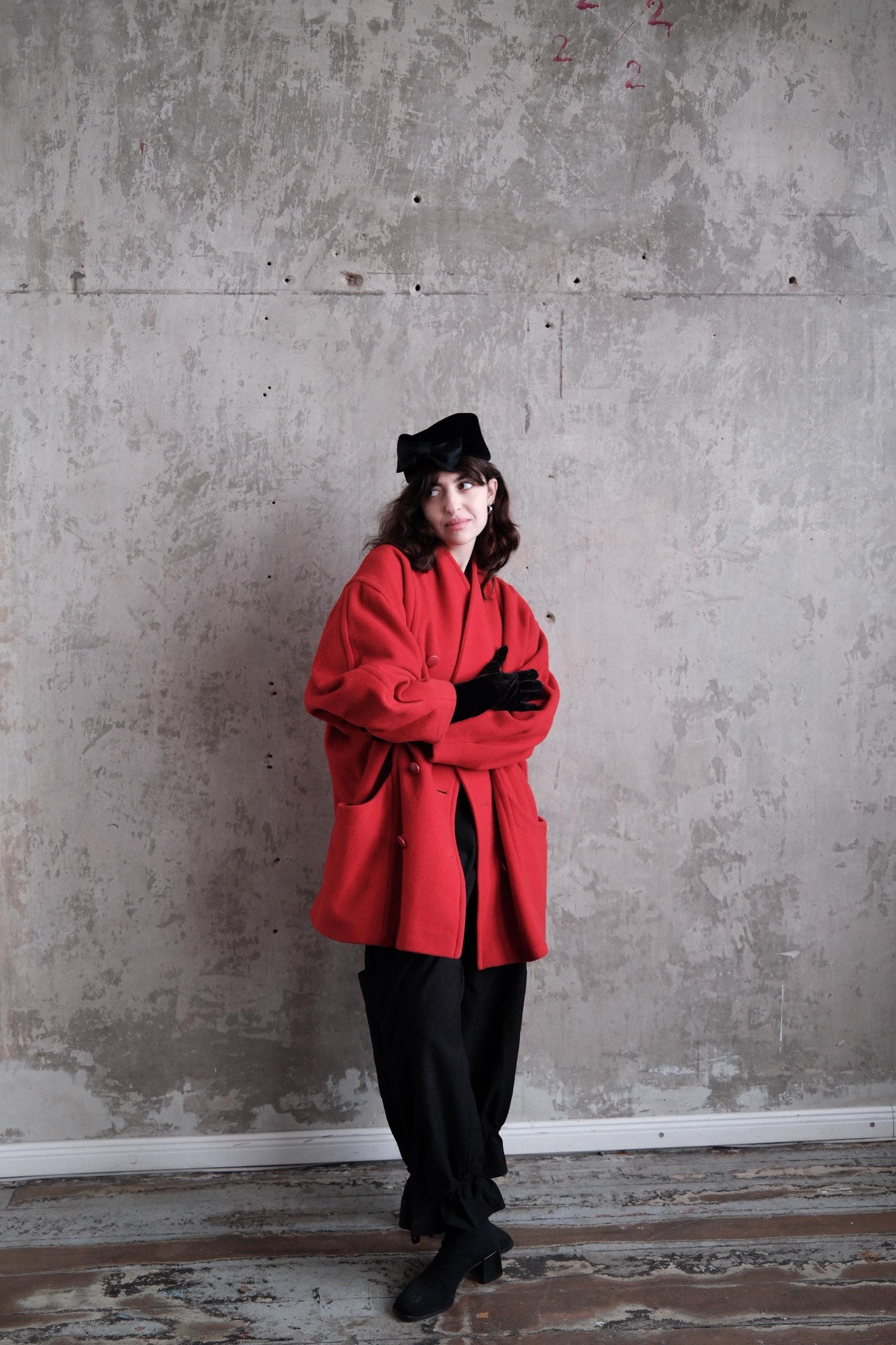 A person wearing a red coat and black pants stands in a room with a concrete wall.
