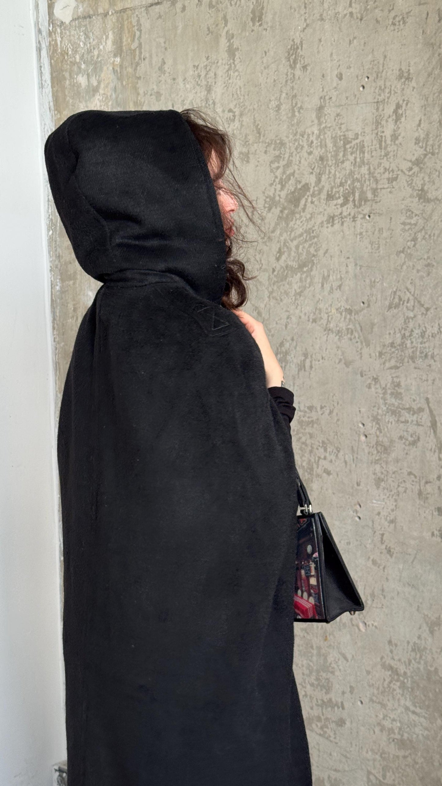 A person wearing a black hooded cloak stands against a concrete wall, holding a black bag.