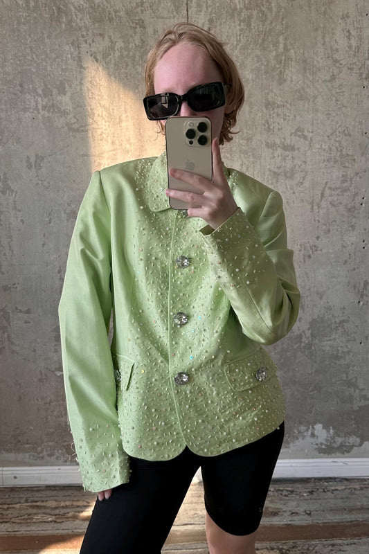 A person wearing a mint green jacket with sparkly details and sunglasses is taking a selfie with a smartphone.