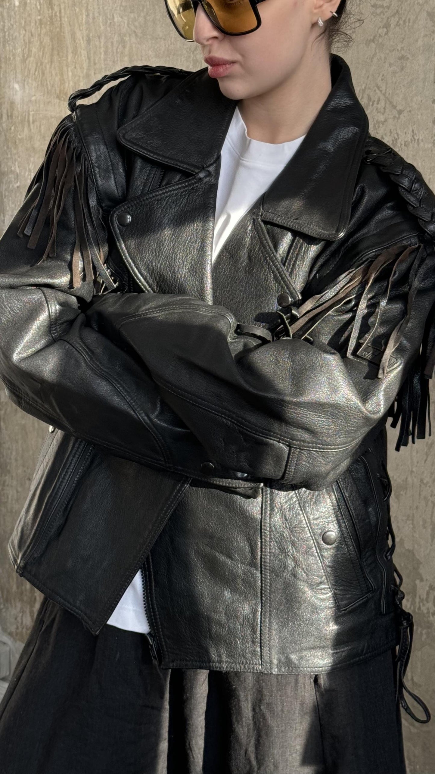 A person wearing a black leather jacket with fringe details, standing against a concrete wall.