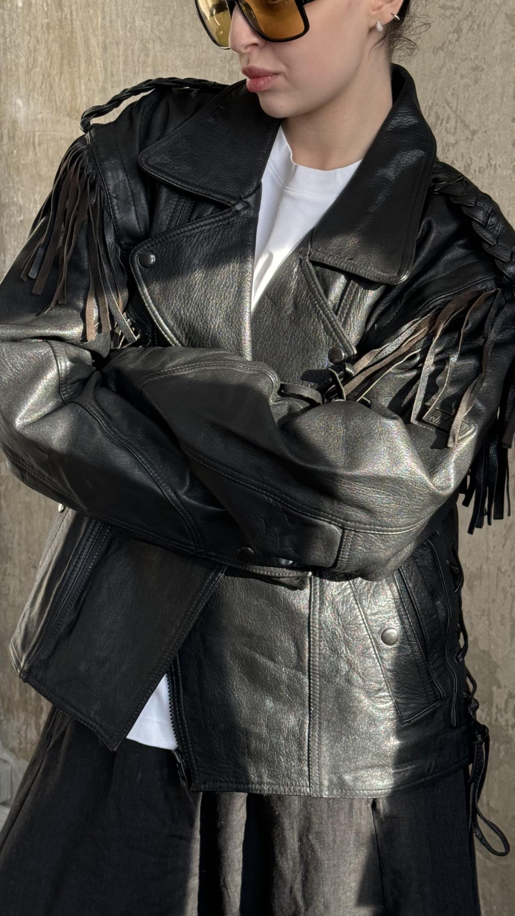 A person wearing a black leather jacket with fringe details, standing against a concrete wall.
