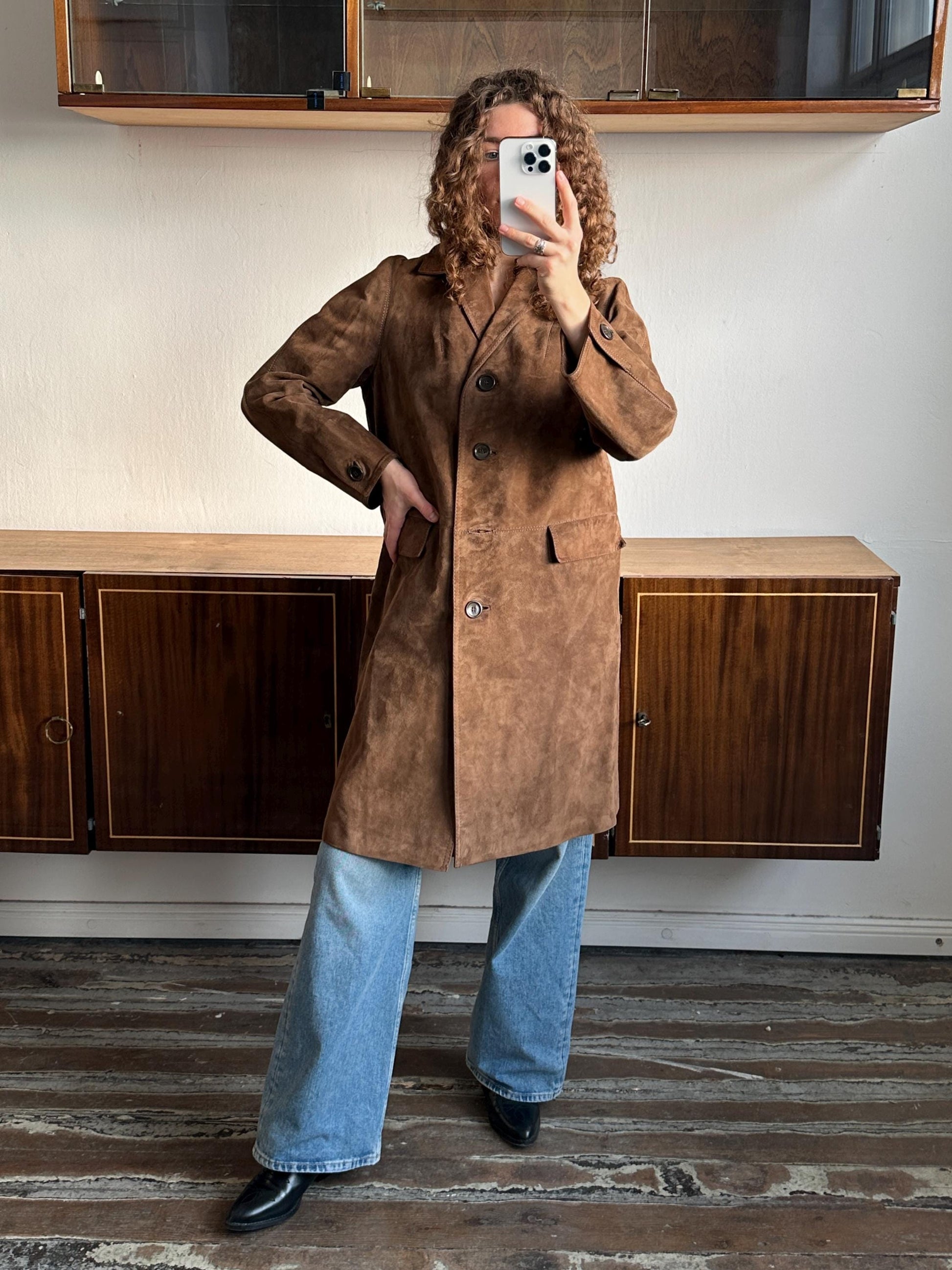 A person is taking a selfie with a brown suede coat and blue jeans, standing in front of a wooden cabinet.