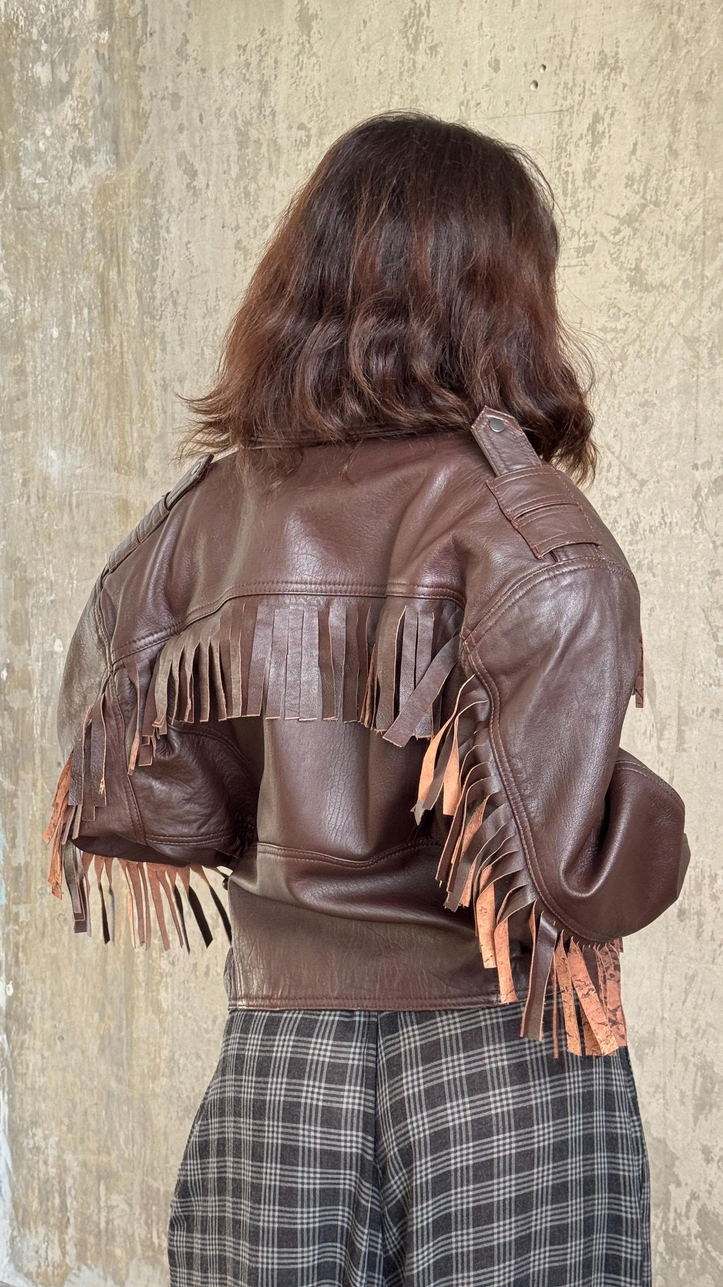 A person wearing a brown leather jacket with fringe details, standing against a concrete wall.