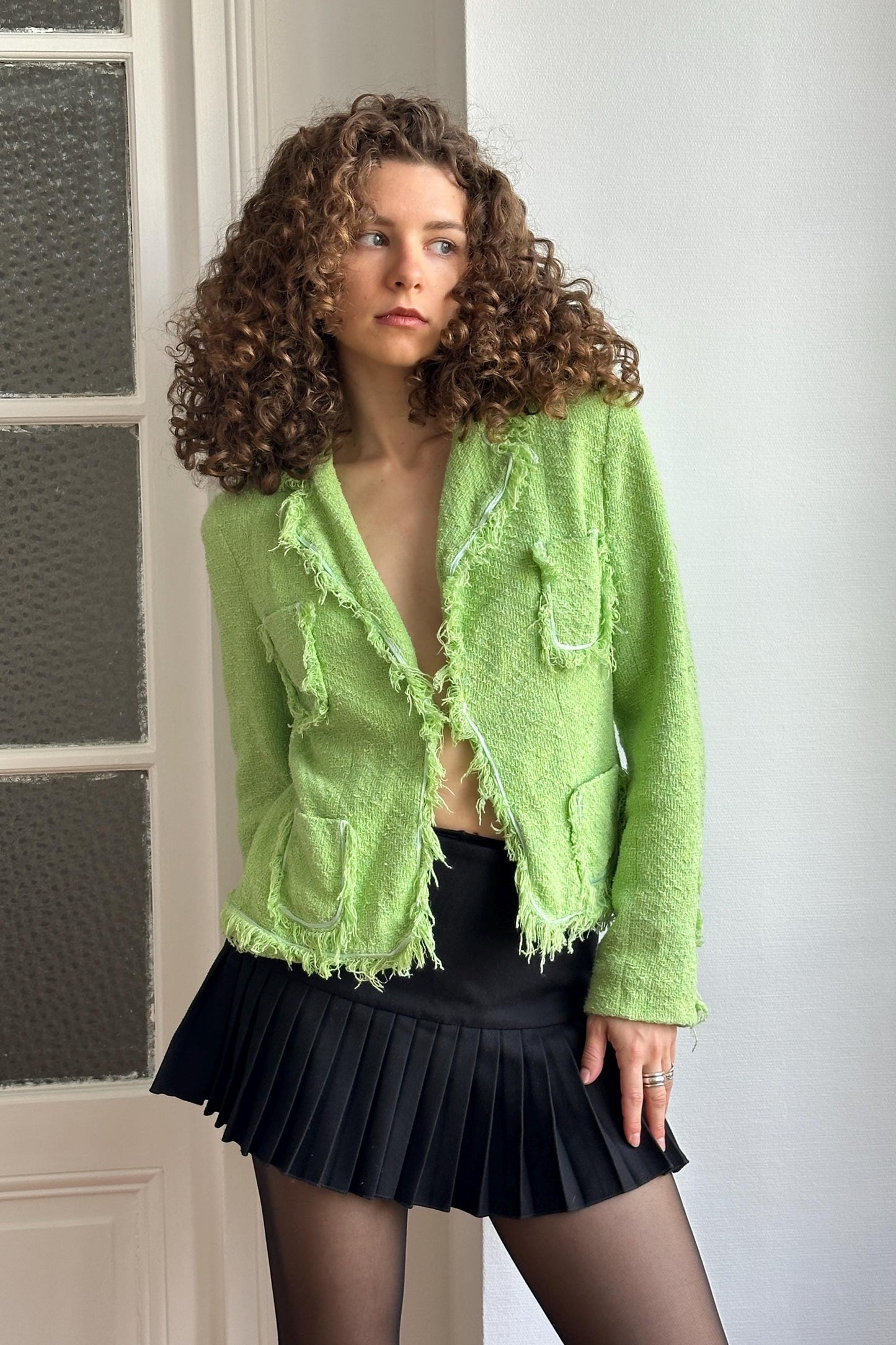 A woman with curly hair wearing a green tweed jacket and a black pleated skirt stands in front of a white door.