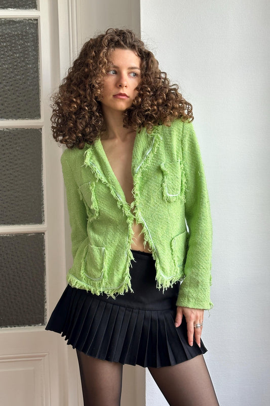 A woman with curly hair wearing a green tweed jacket and a black pleated skirt stands in front of a white door.