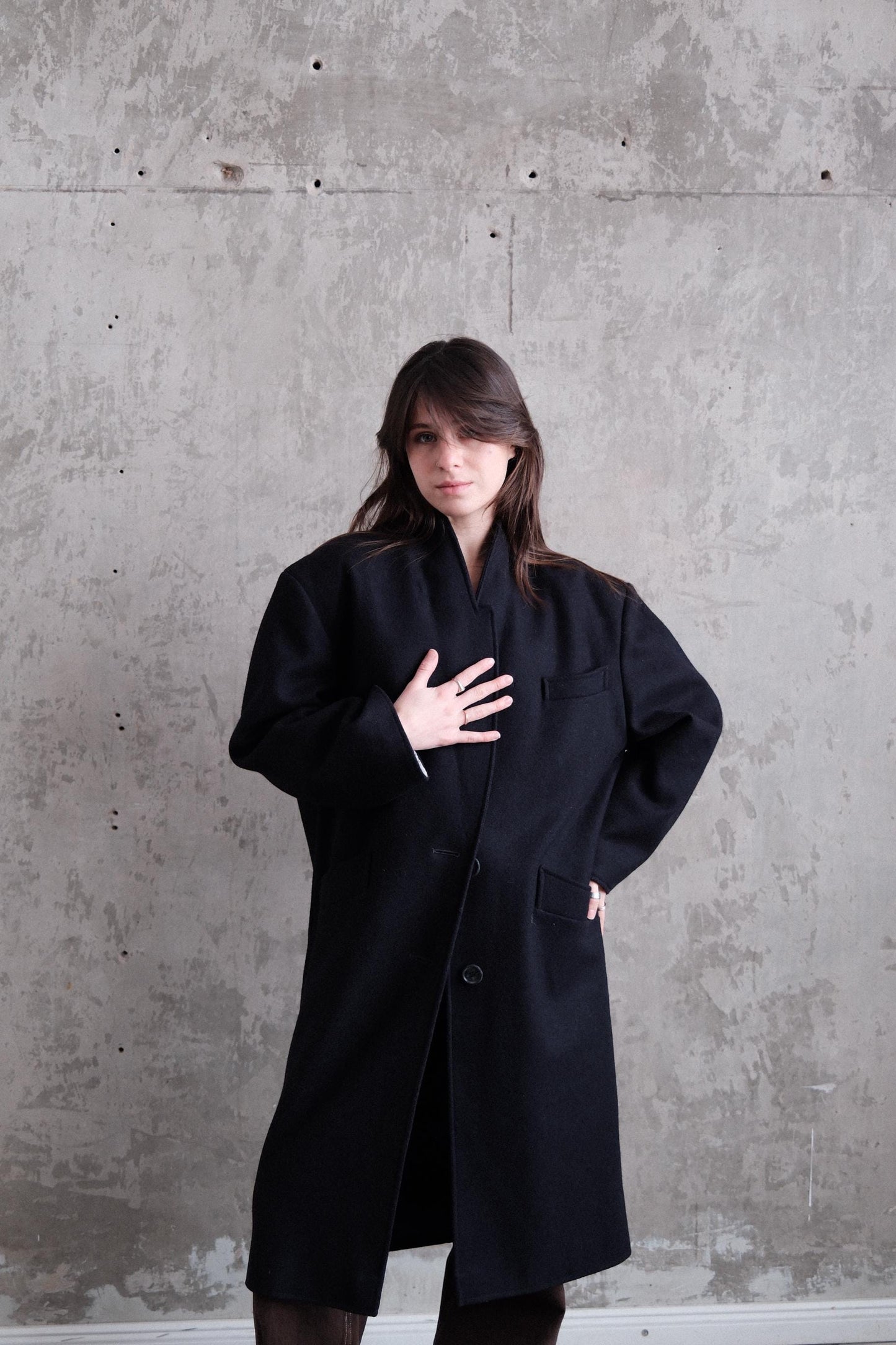 A woman in a black coat stands against a concrete wall, her gaze directed downwards.