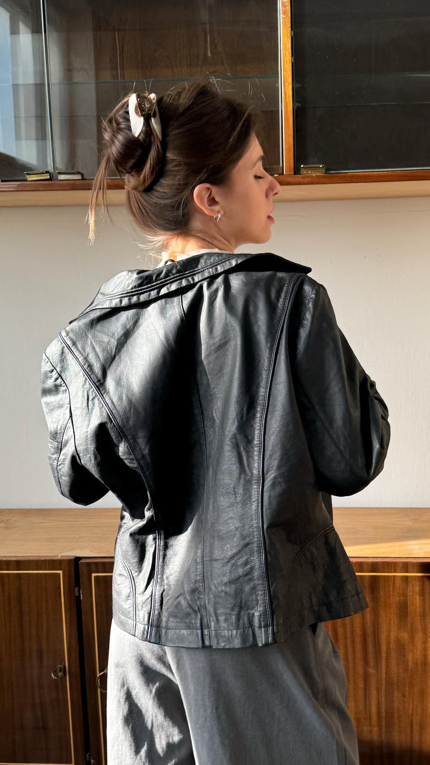 A woman wearing a black leather jacket stands in front of a wooden cabinet, her back facing the camera.