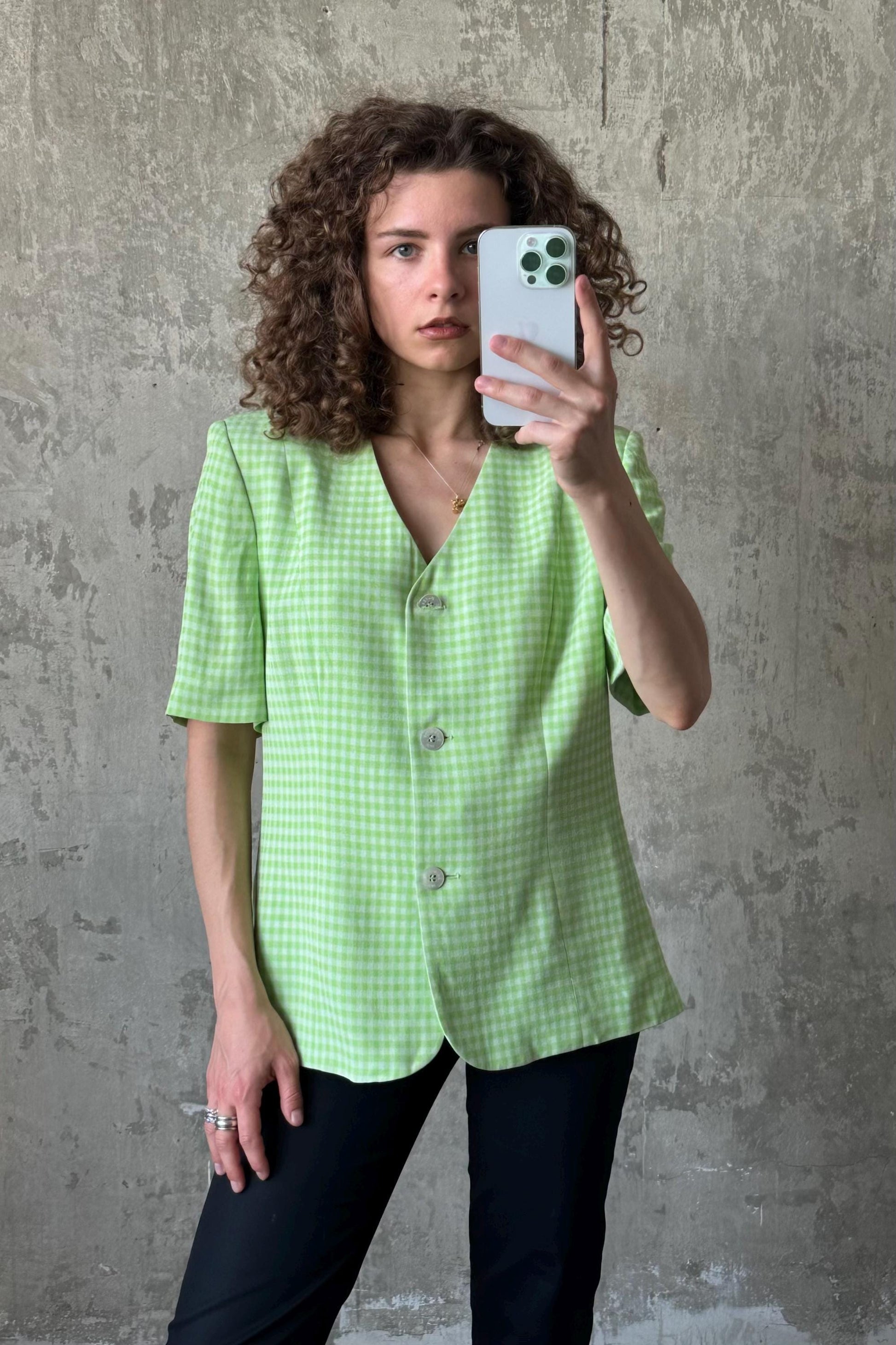 A woman with curly hair is taking a selfie with a white iPhone, wearing a green and white checkered blazer.
