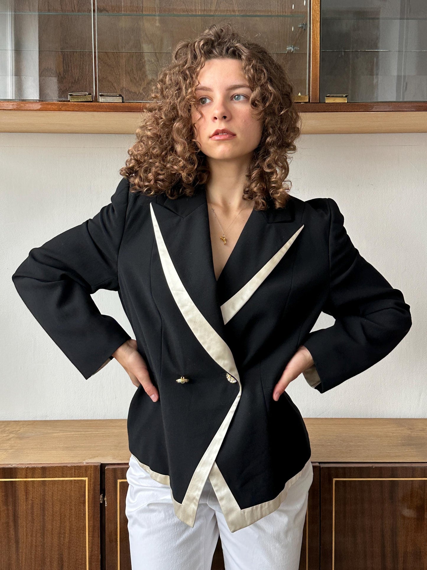 A woman with curly hair wearing a black blazer with white accents stands in front of a wooden cabinet.