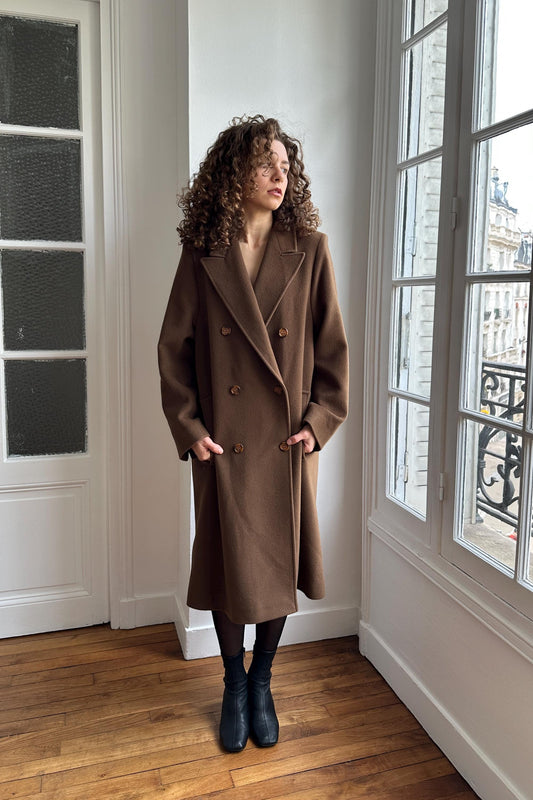 A woman with curly hair stands in a room, wearing a brown coat and black boots.