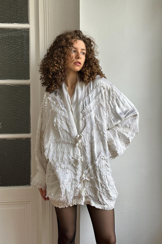 A woman with curly hair wearing a white, ruffled jacket stands in front of a white door.