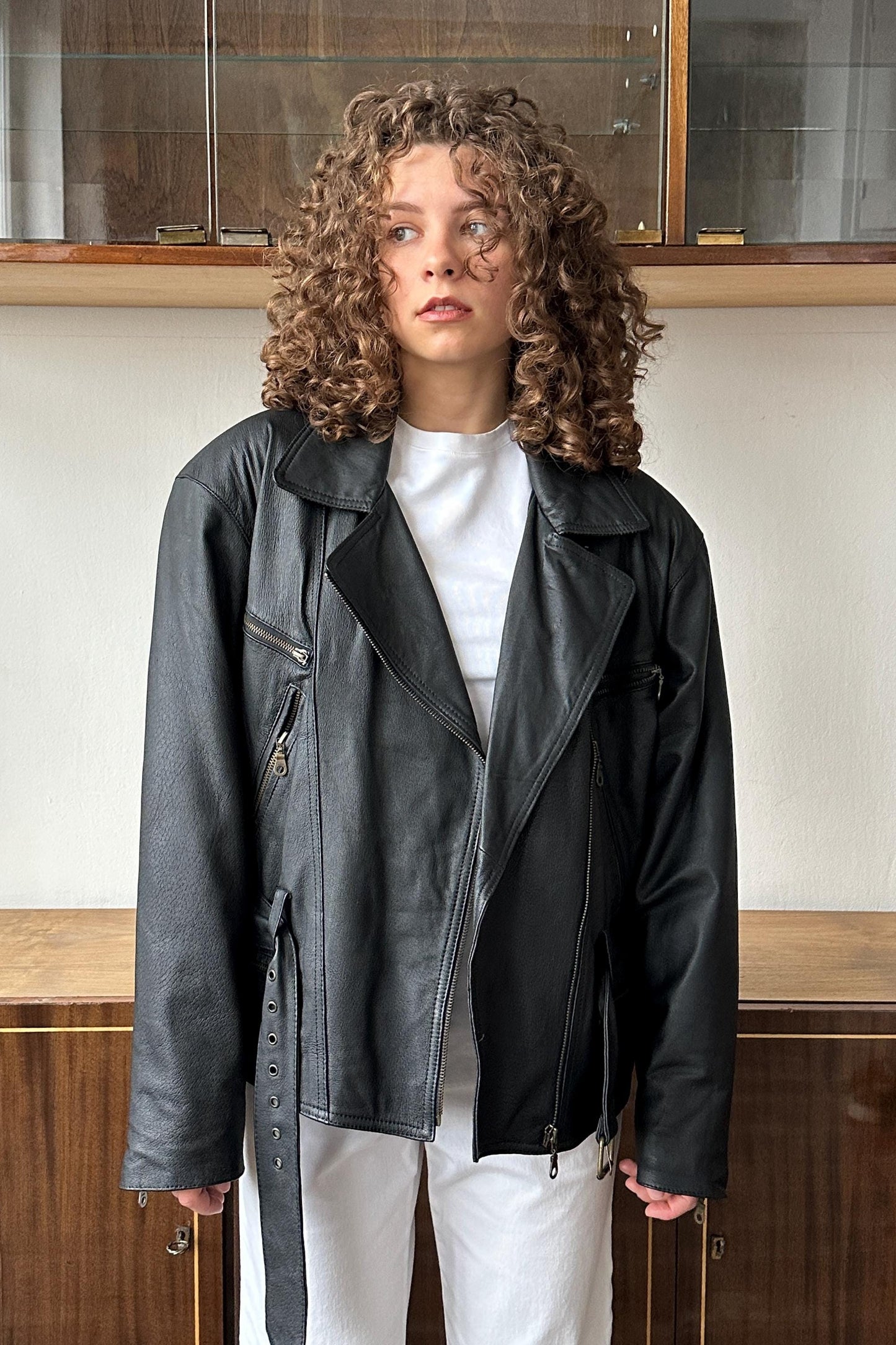 A woman with curly hair wearing a black leather jacket stands in front of a glass display case.