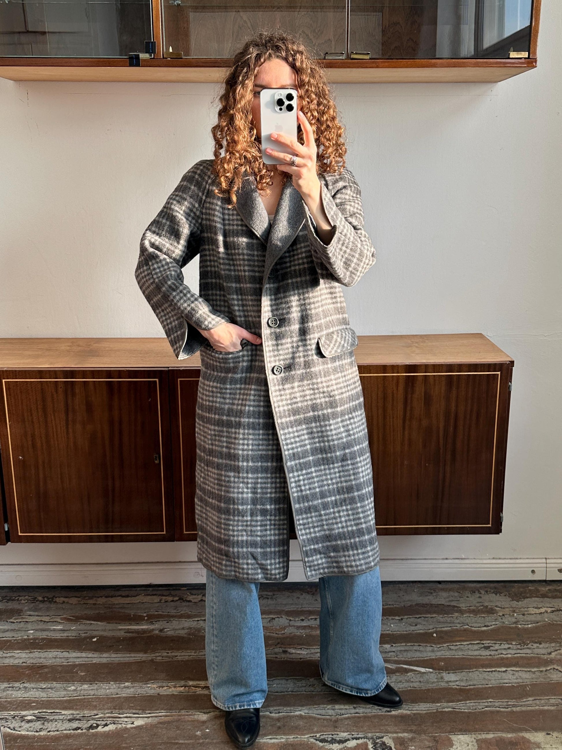 A person with curly hair is taking a selfie with a smartphone, wearing a gray plaid coat and blue jeans.