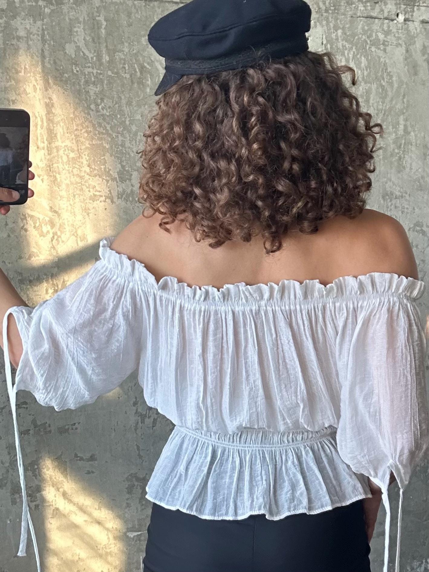 a person taking a selfie with a smartphone, wearing a white off-the-shoulder blouse and a black cap.