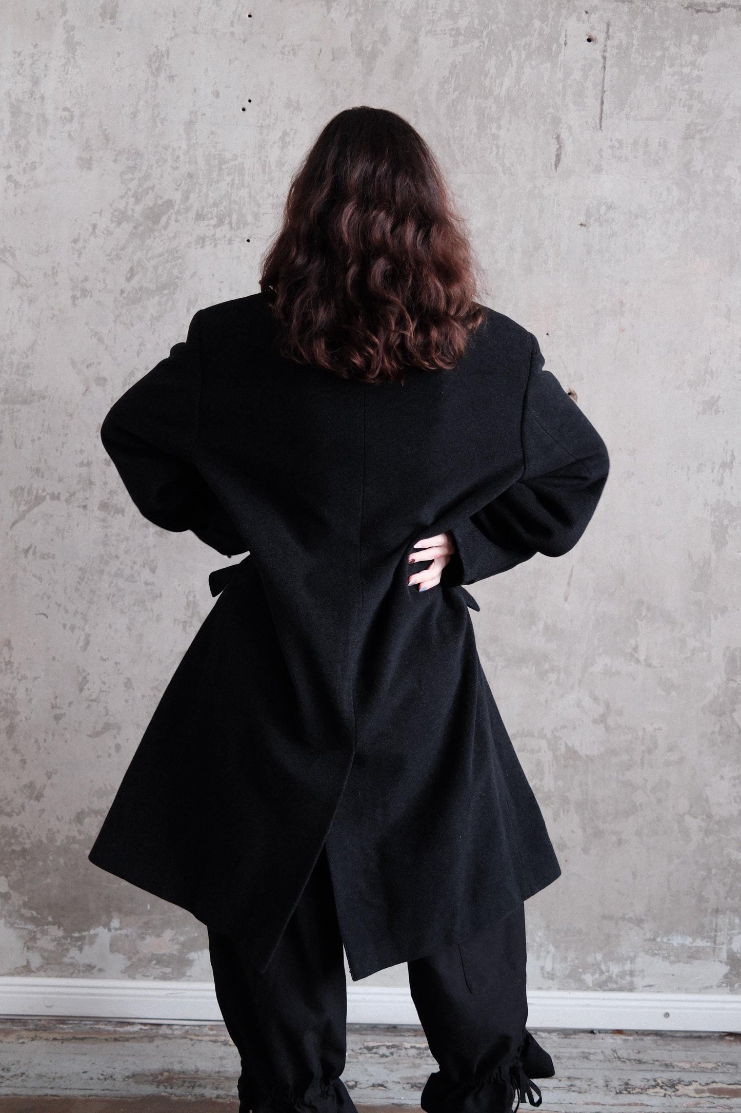 A person wearing a black coat stands against a plain white wall.