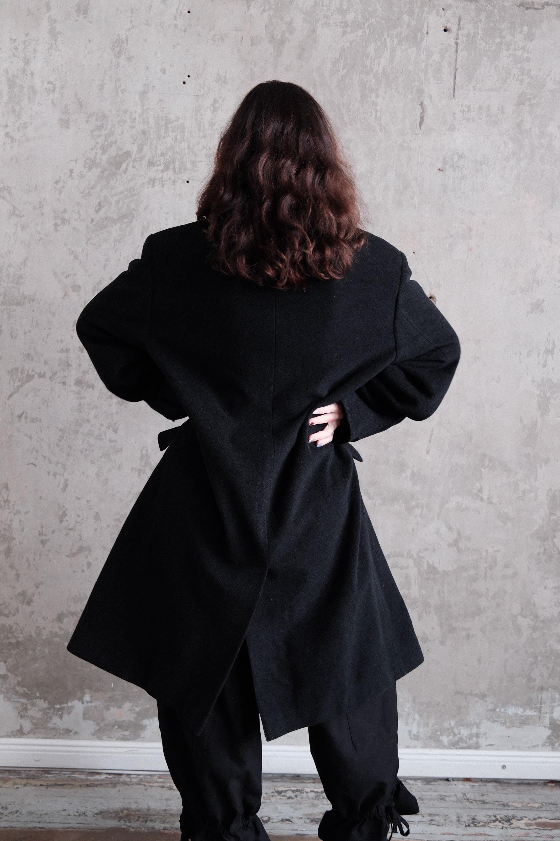 A person wearing a black coat stands against a plain white wall.
