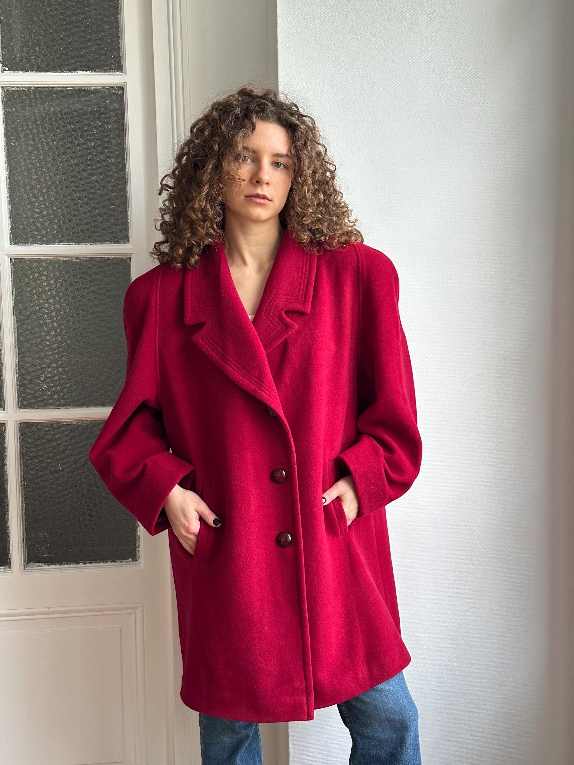 A person wearing a red coat is leaning against a white wall, with their head tilted back and hands in their pockets.