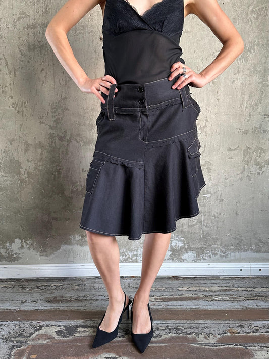A woman wearing a black skirt and top stands confidently with her hands on her hips against a concrete wall.
