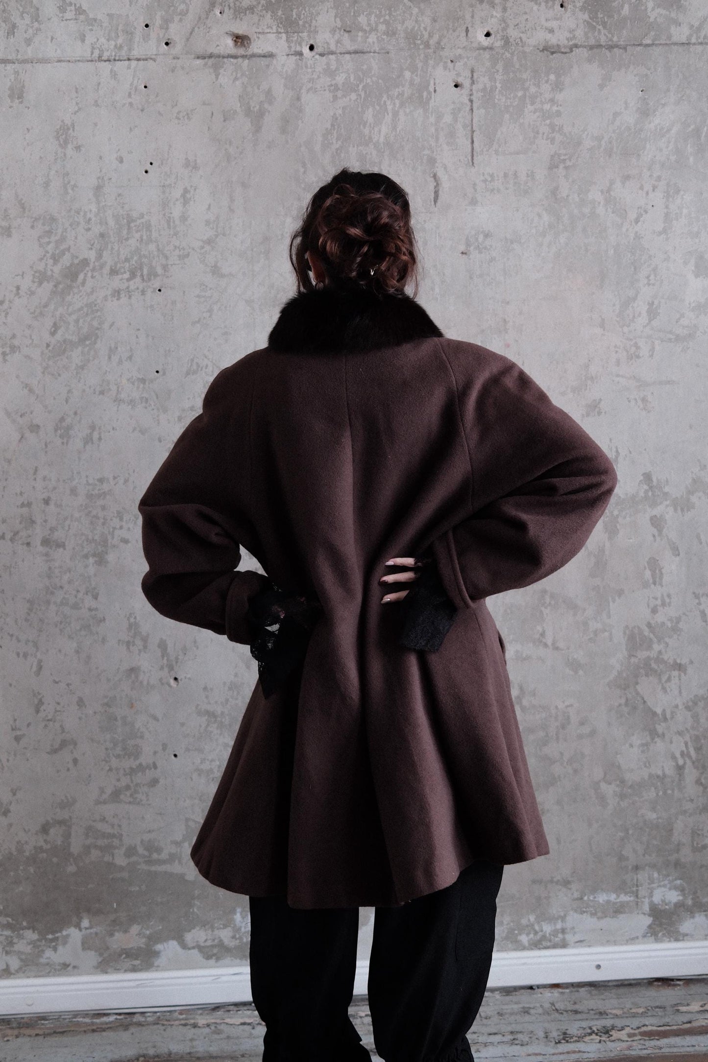 a person wearing a brown coat standing against a concrete wall.