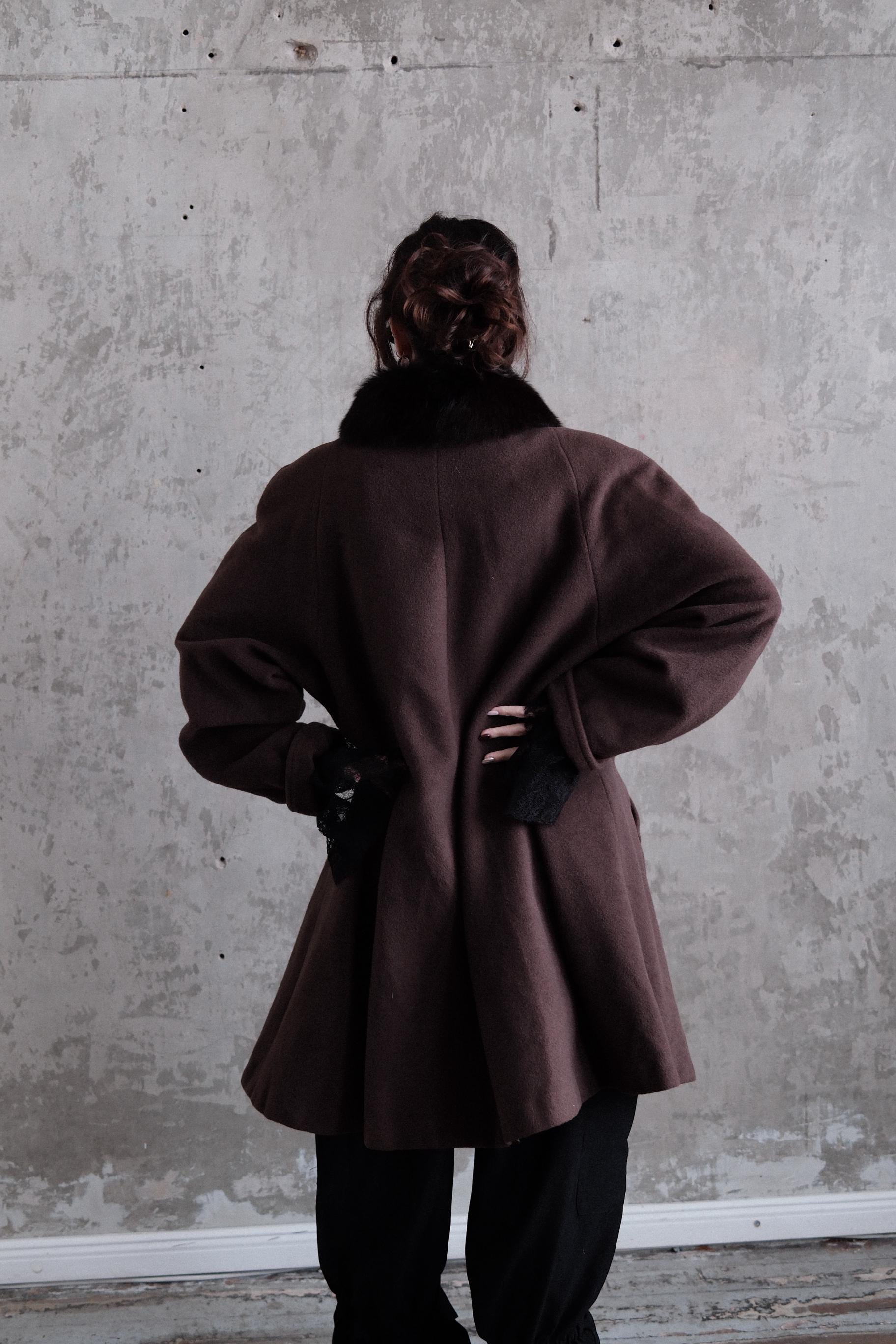 a person wearing a brown coat standing against a concrete wall.
