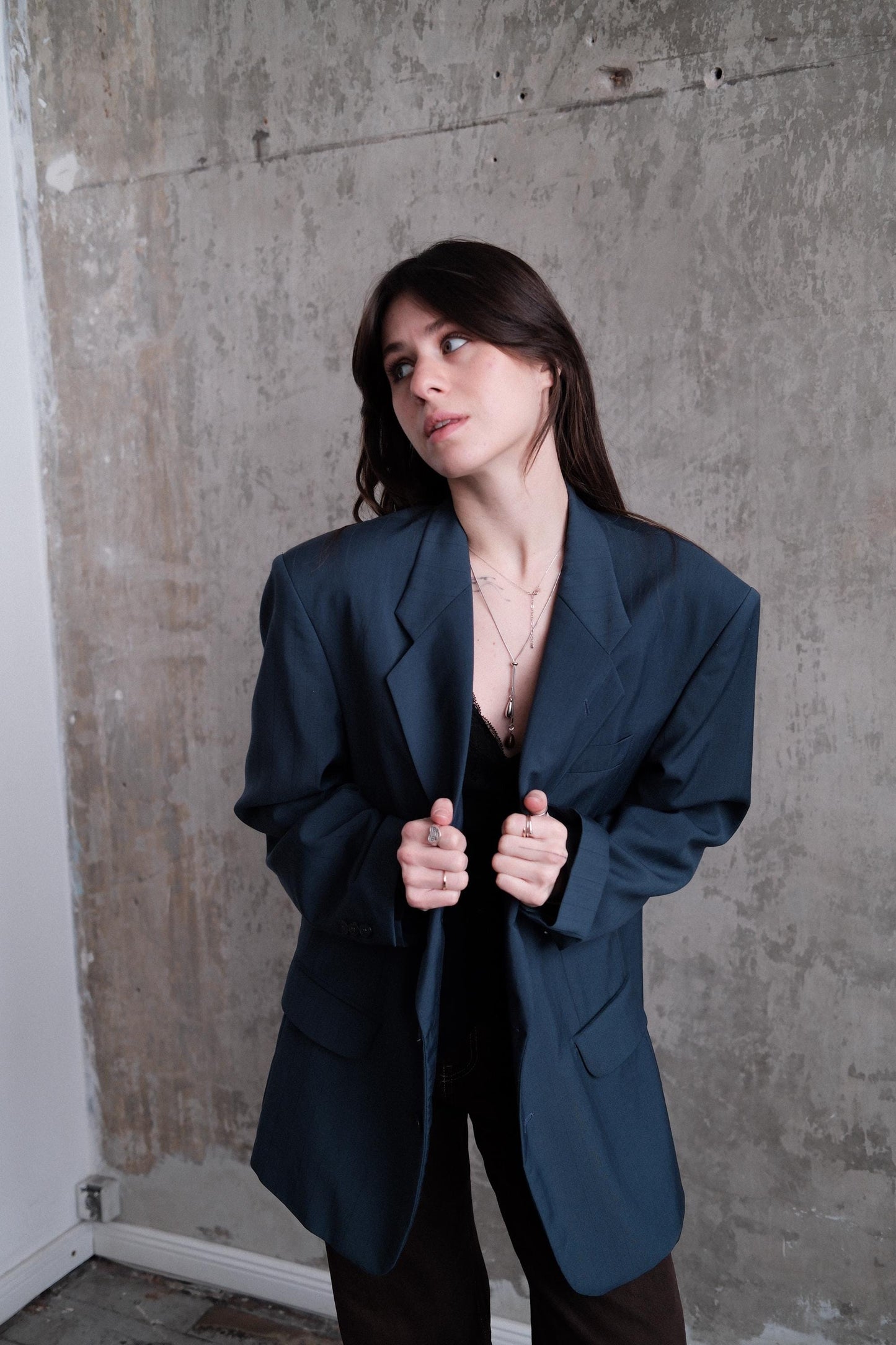 A woman in a dark blue blazer stands against a concrete wall, her hands clasped behind her back.