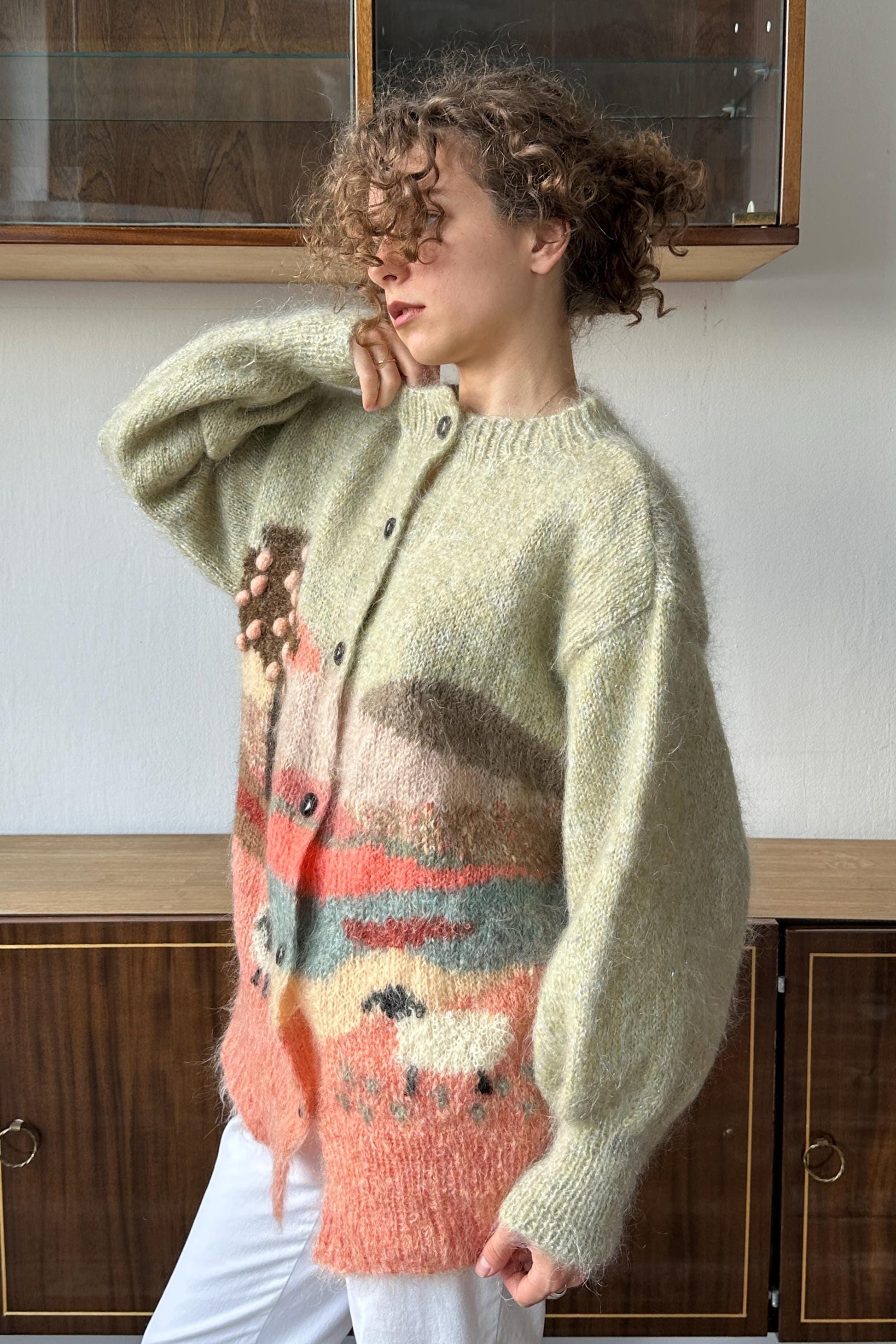 A woman wearing a light pink and orange knitted cardigan stands in front of a wooden cabinet.