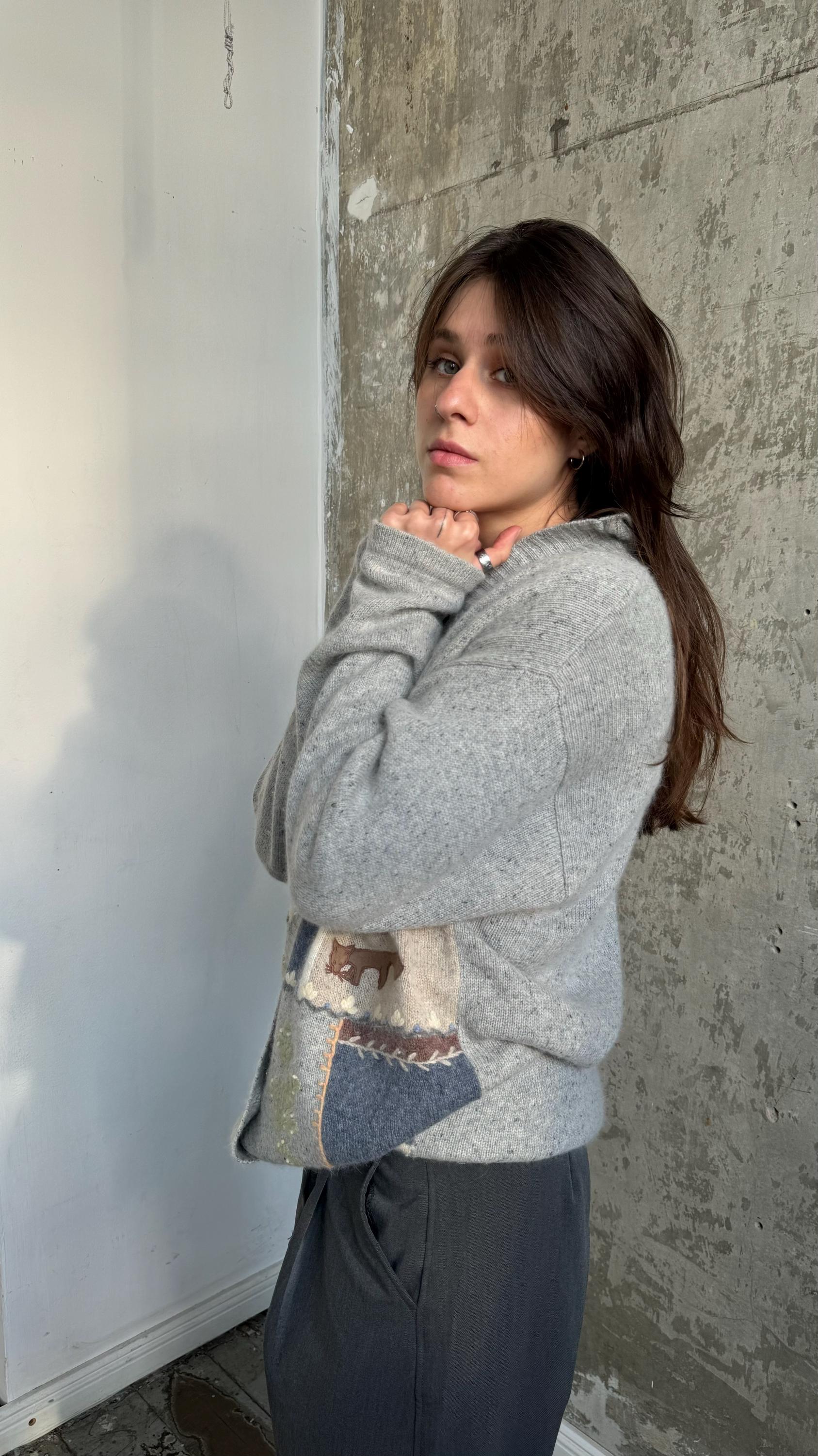 A young woman with long dark hair stands in front of a concrete wall, wearing a gray sweater and dark pants. She has her hands tucked into her pockets and is looking off to the side.