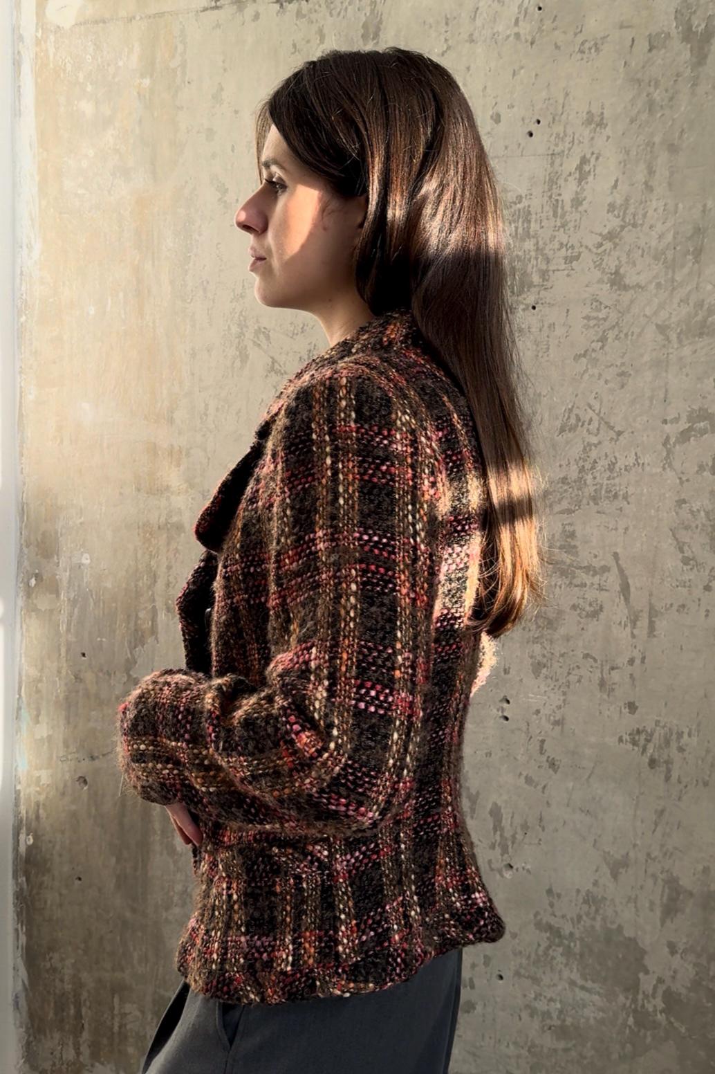 A woman with long brown hair wearing a brown and black plaid jacket stands against a concrete wall, looking to the side.