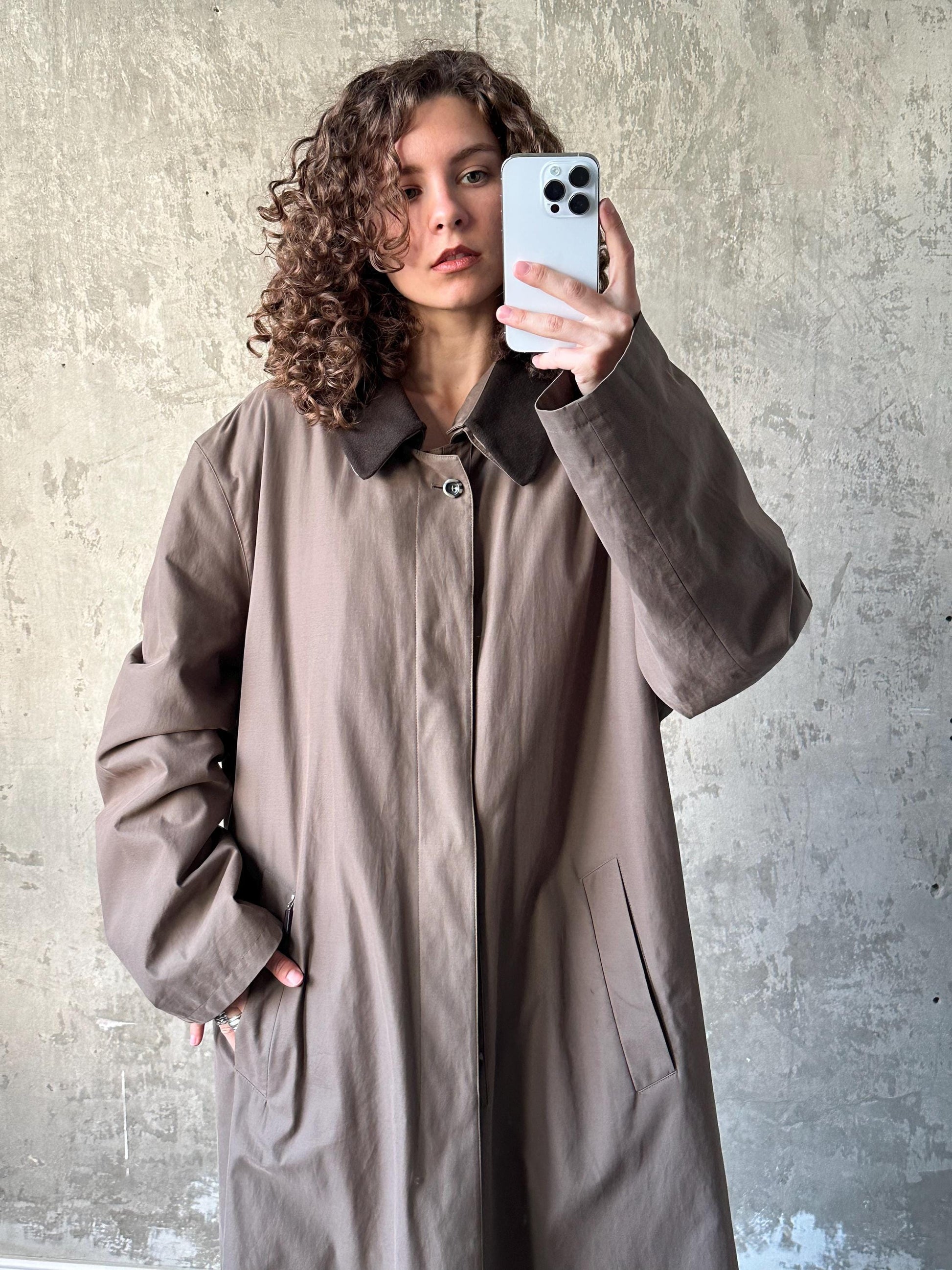 A woman with curly hair is taking a selfie with a white smartphone, wearing a beige coat.