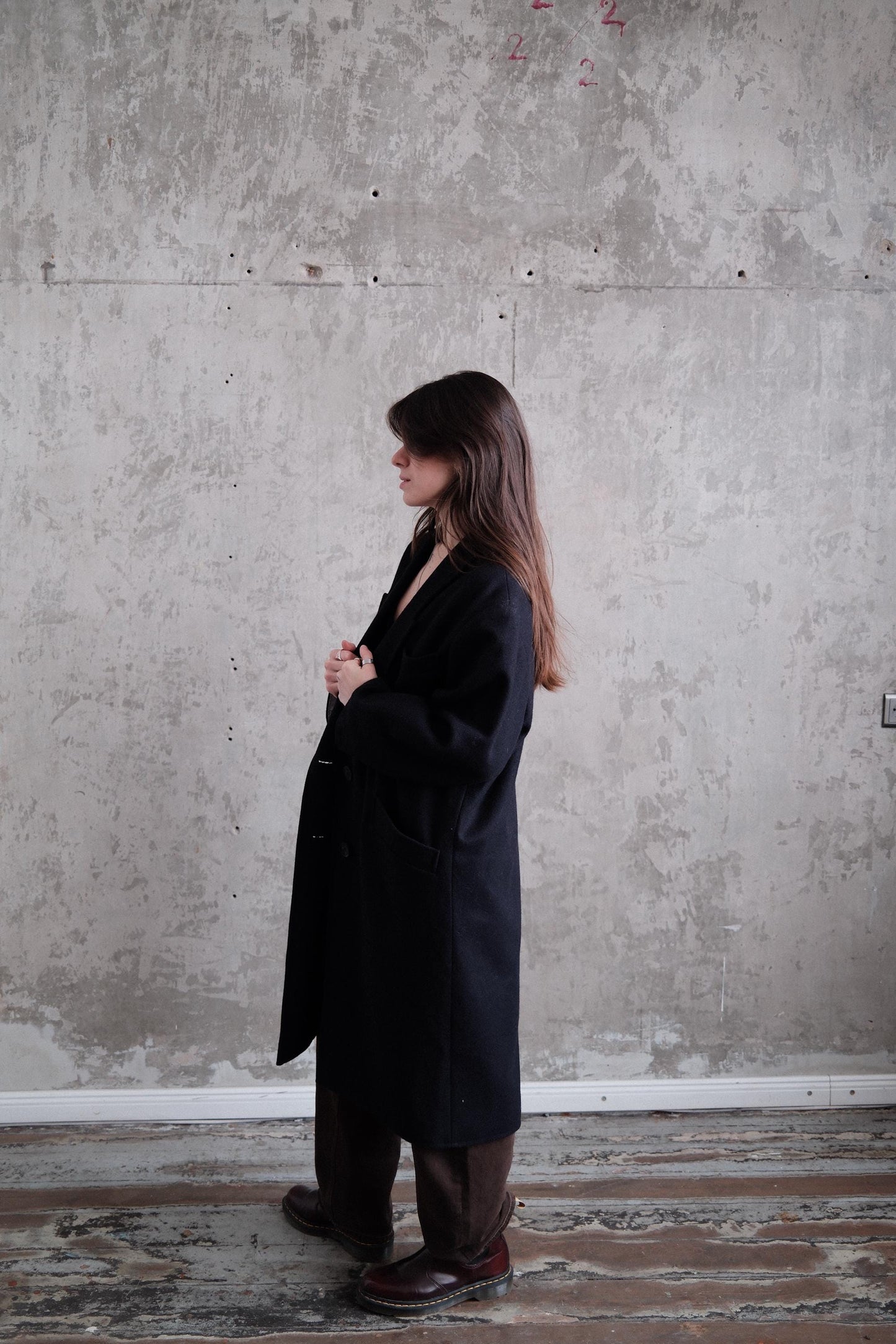 A person wearing a black coat stands in a room with a concrete wall.