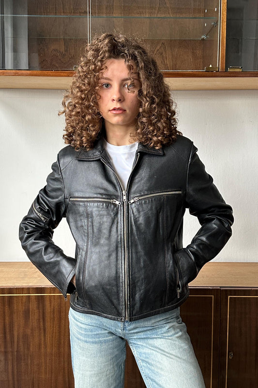 A young woman with curly hair wearing a black leather jacket stands in front of a wooden cabinet.