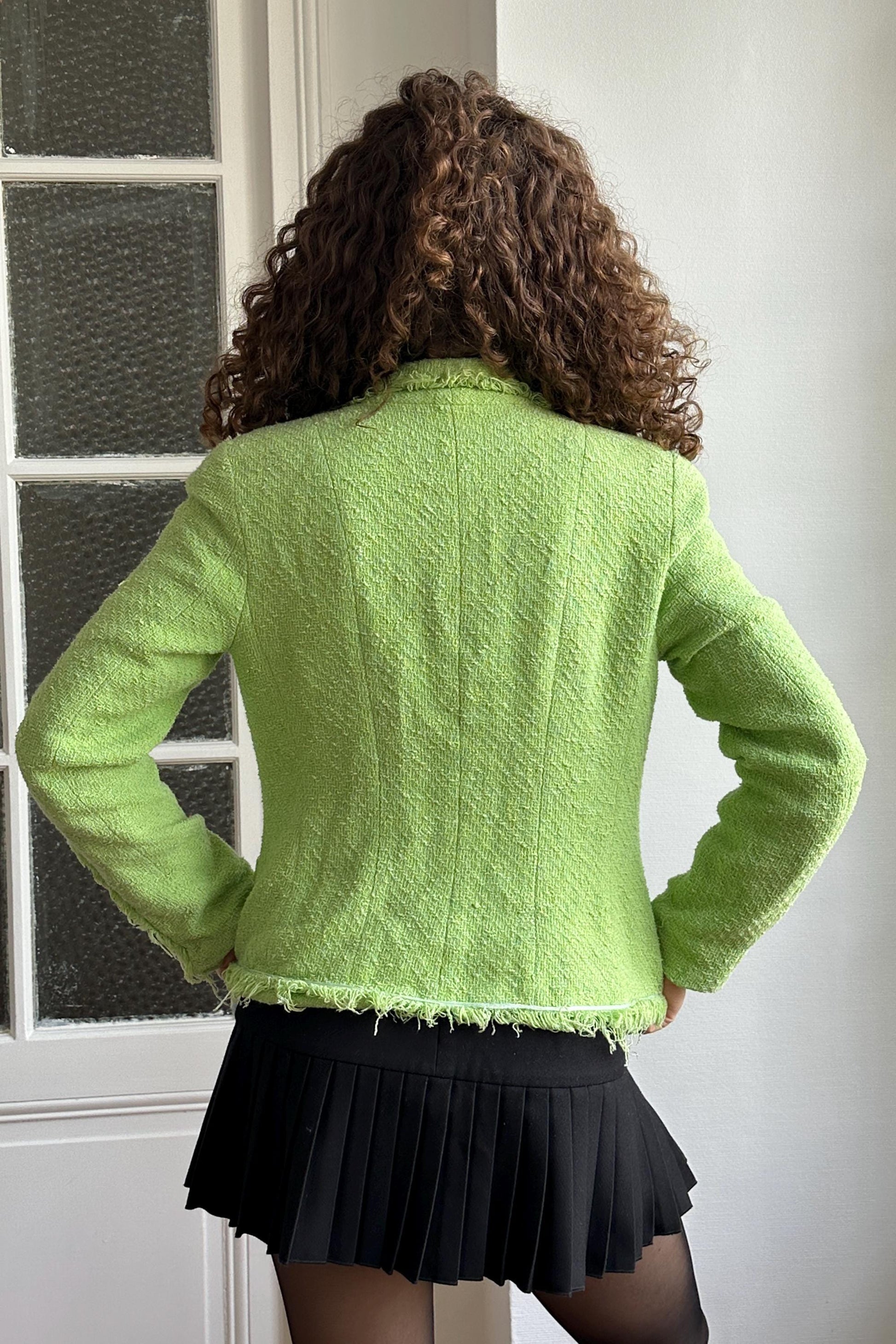 A woman with curly hair wearing a lime green tweed jacket and a black pleated skirt stands in front of a window.