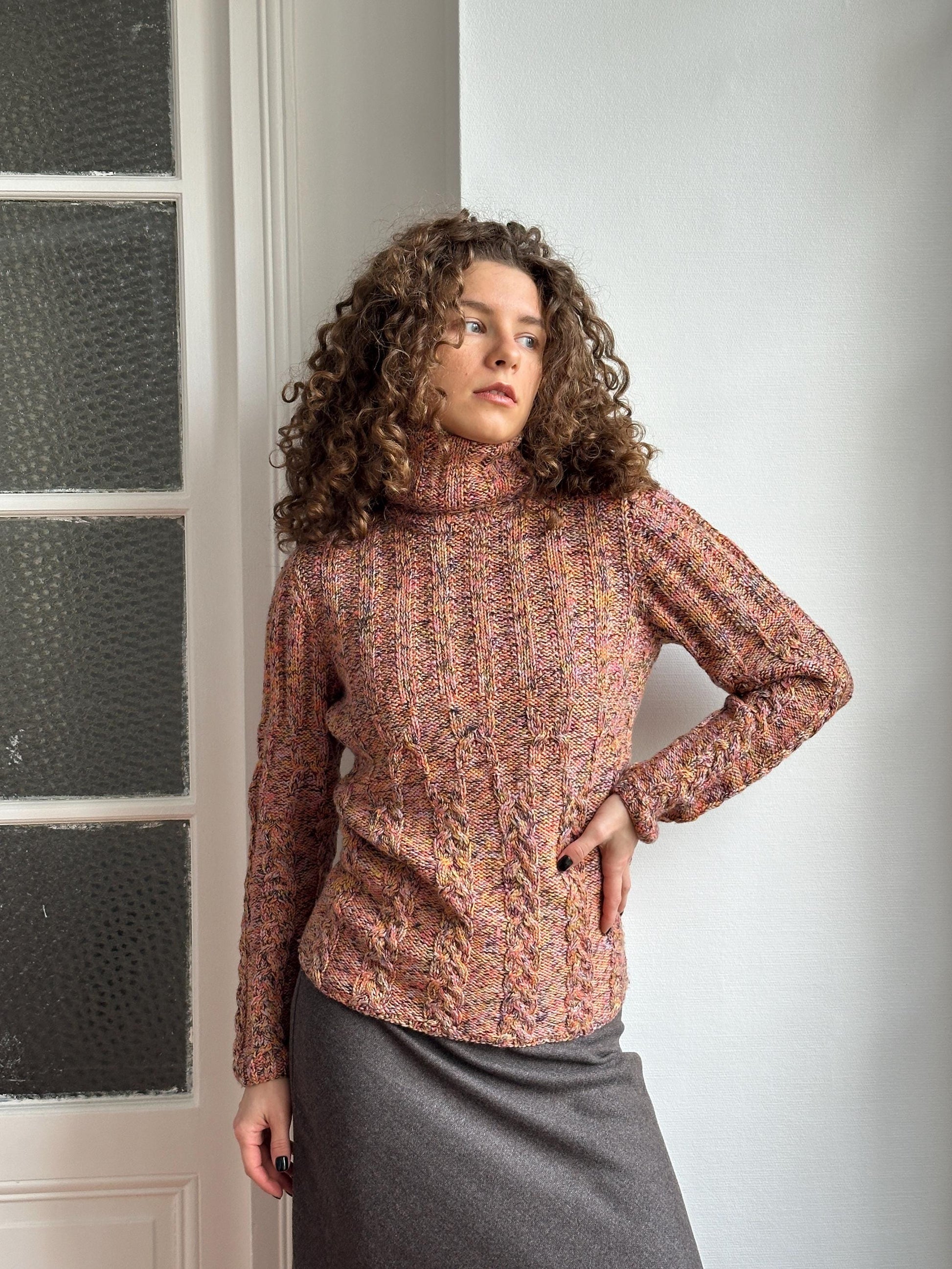 A woman with curly hair wearing a colorful sweater is leaning against a white wall, with her hand on her hip.