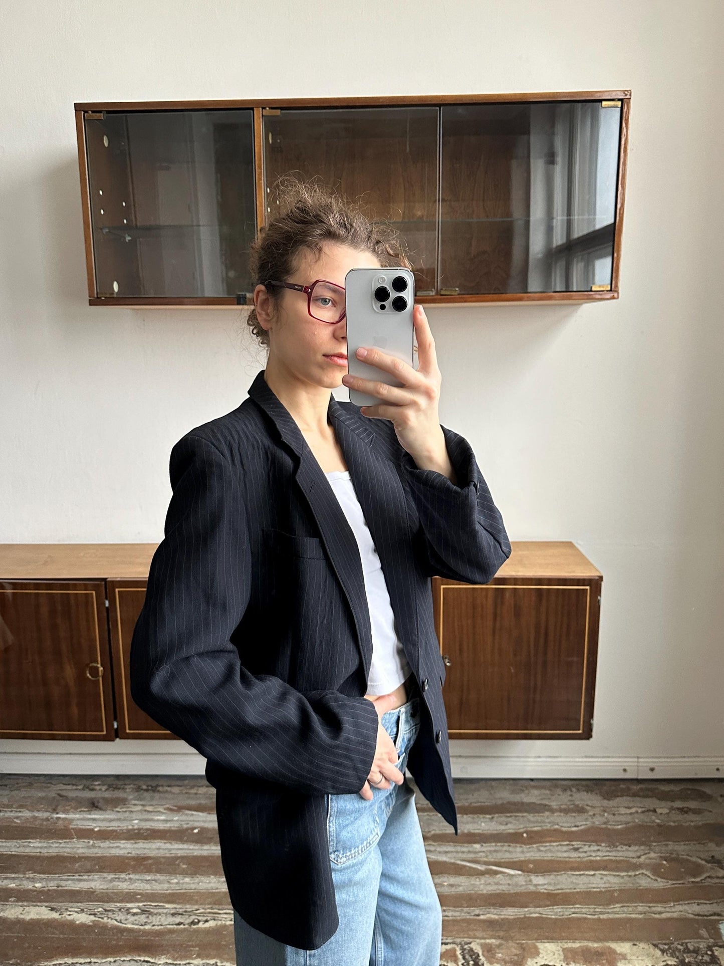 A woman wearing a black blazer and jeans is taking a selfie with a smartphone in front of a wooden cabinet.