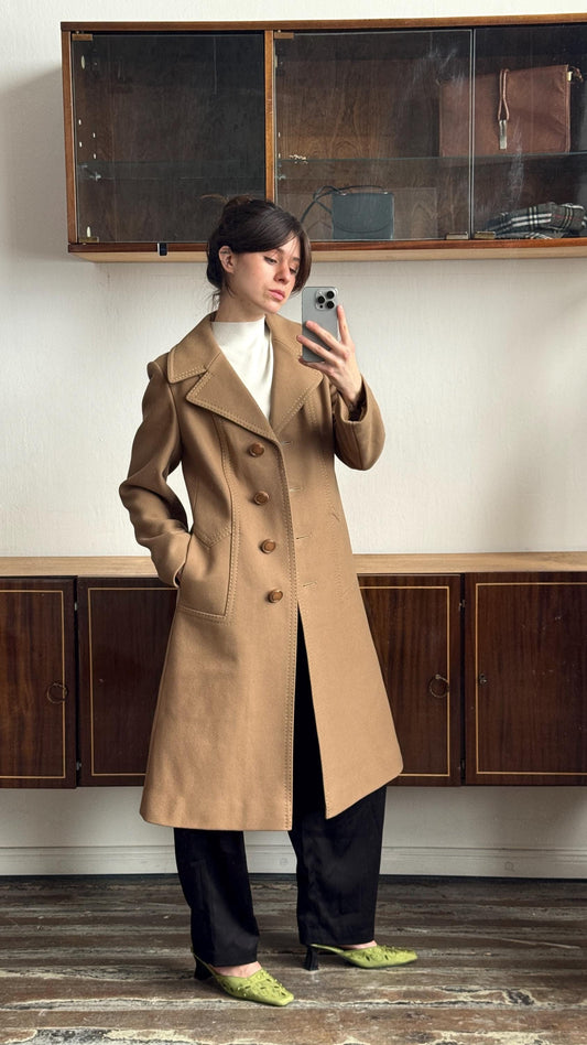 May include: A tan, knee-length coat with a tailored fit. The coat features a notched collar, button closure, and front pockets. The coat is worn over a white top and black trousers. The coat is approximately 114 cm long.