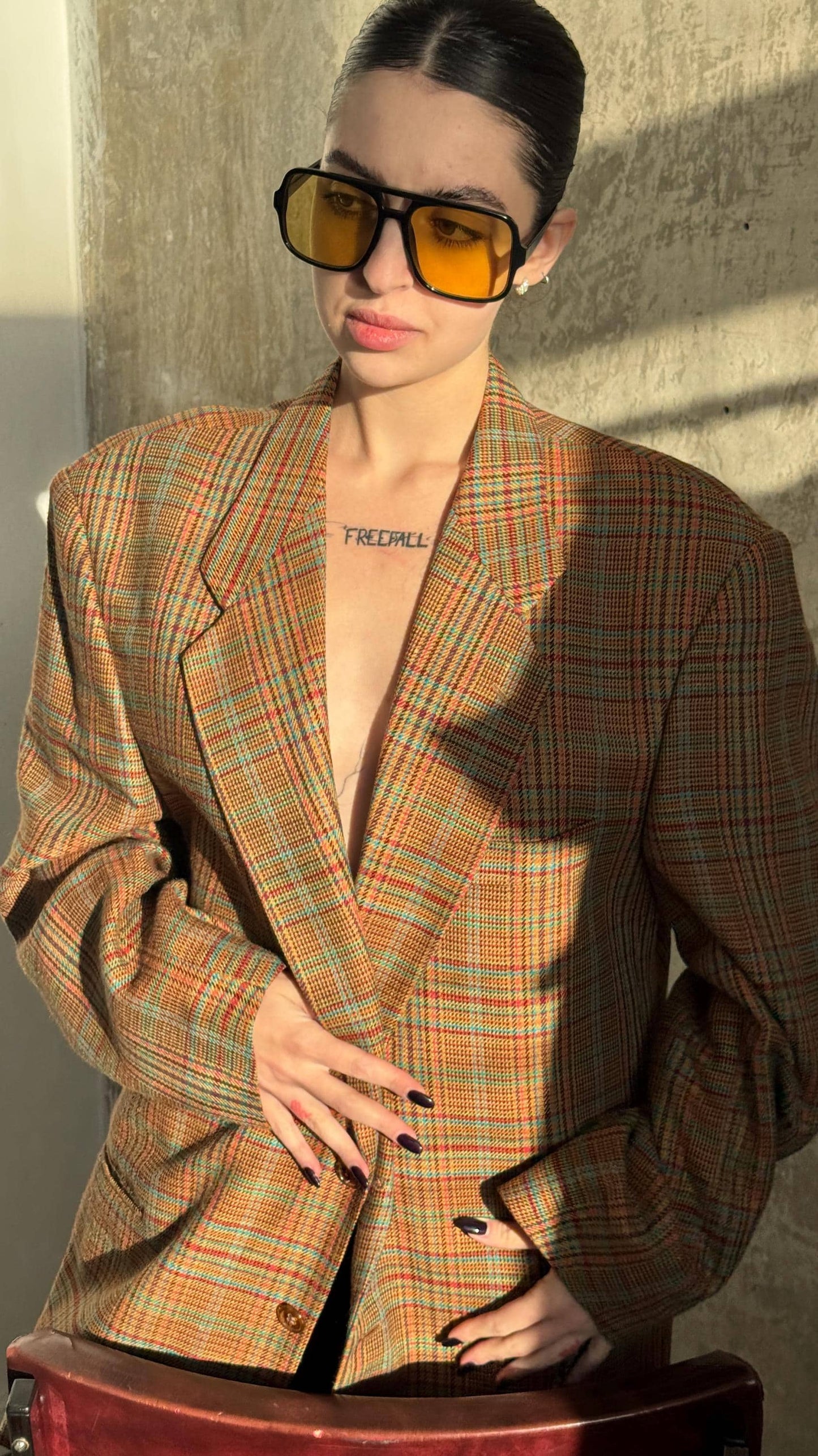 A woman wearing a plaid jacket, sunglasses, and a necklace stands in front of a concrete wall, with her hands on her hips.