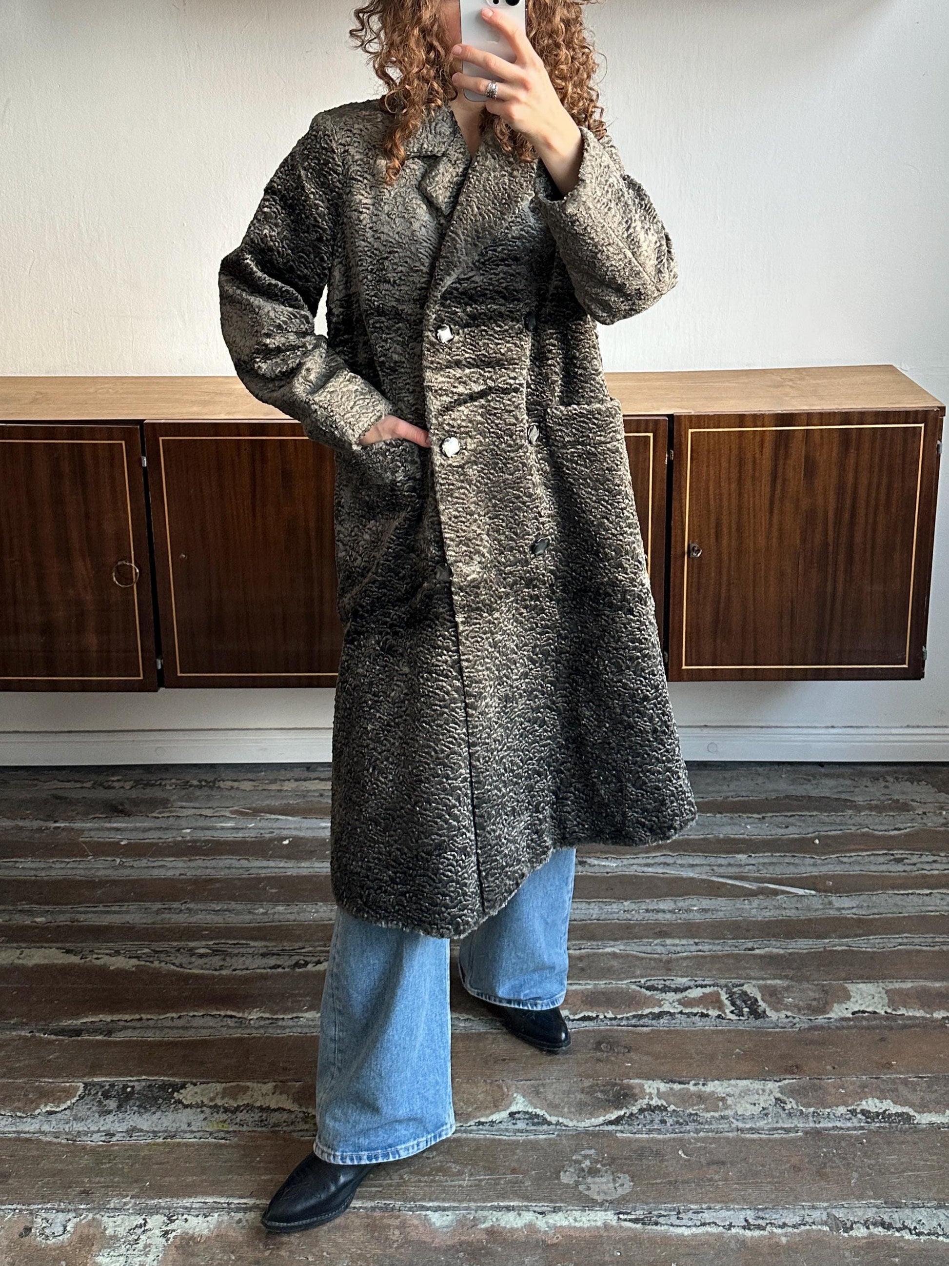 A person is taking a selfie with a gray coat and blue jeans, standing in front of a wooden cabinet.